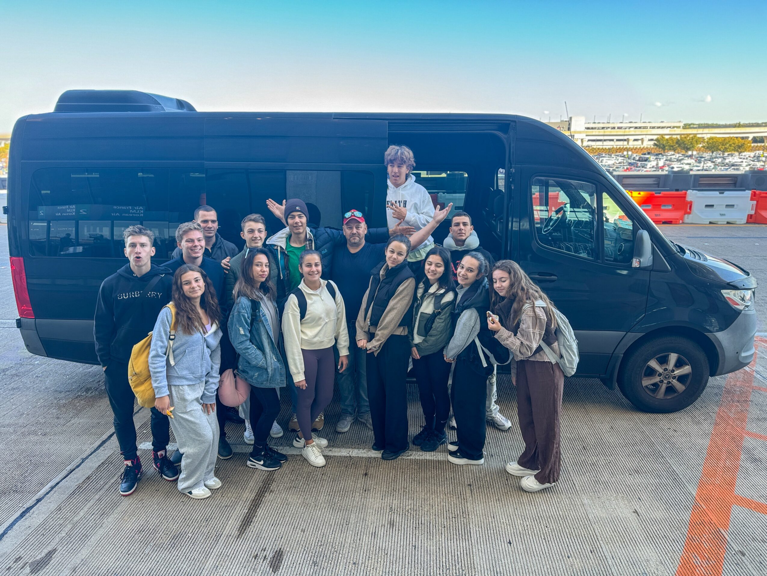 Group of people standing in front of a black van outdoors, with a clear sky and distant buildings in background.