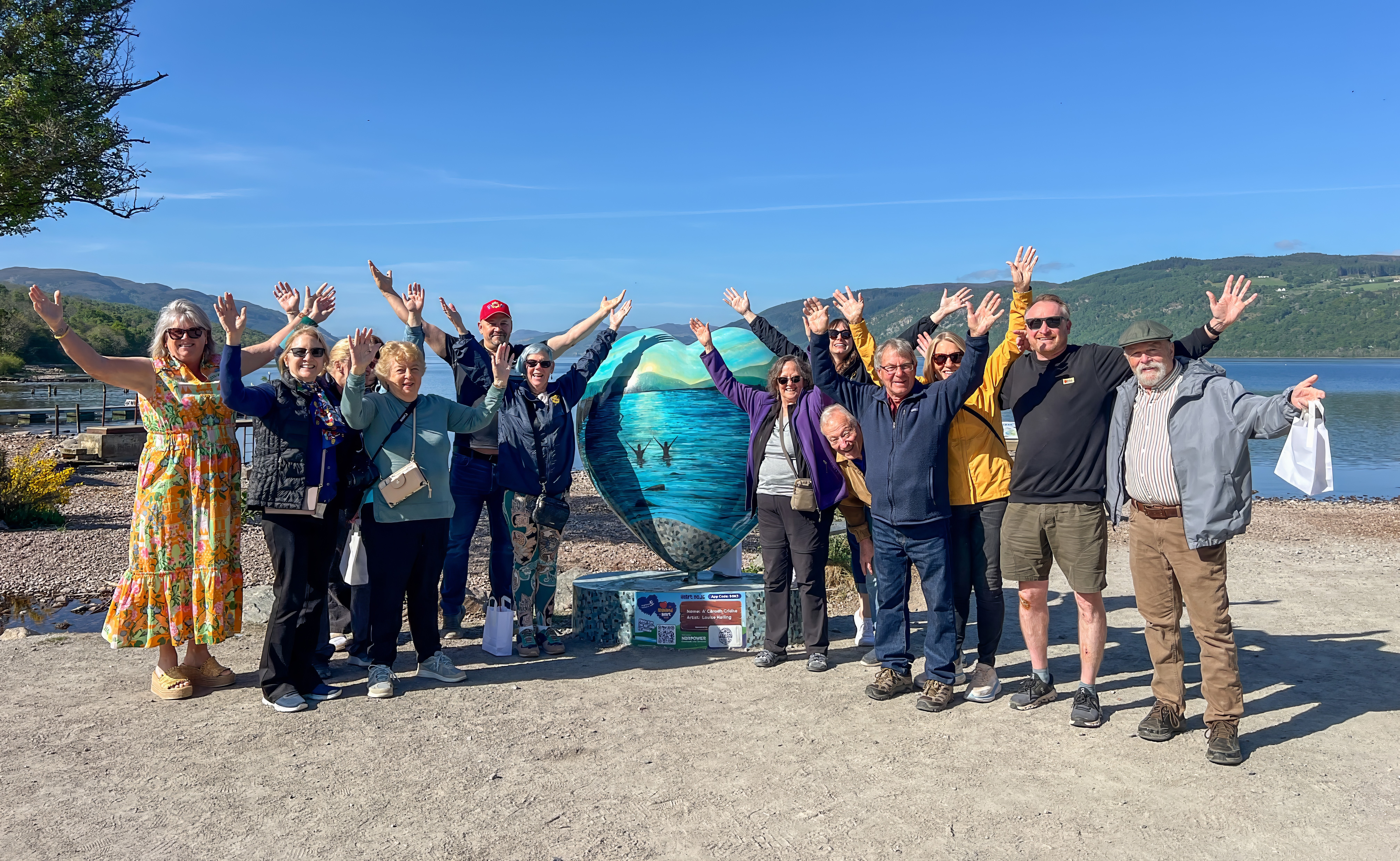 Group of people standing outdoors with arms raised, in front of a large globe sculpture, clear sky, mountains in background.