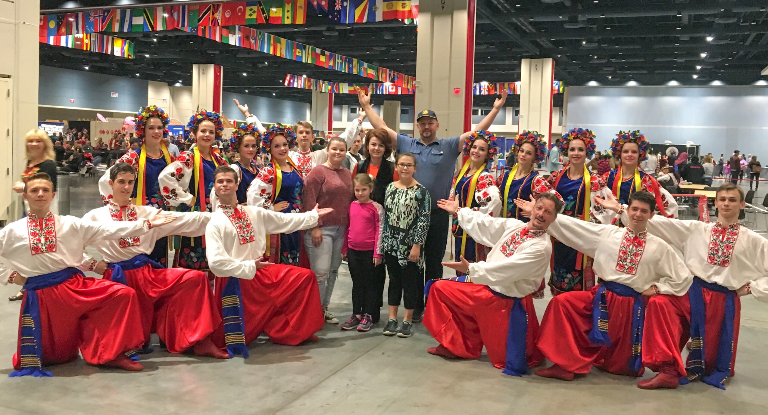 Group of dancers in traditional costumes with arms outstretched, standing in a large indoor event space with colorful decorations.