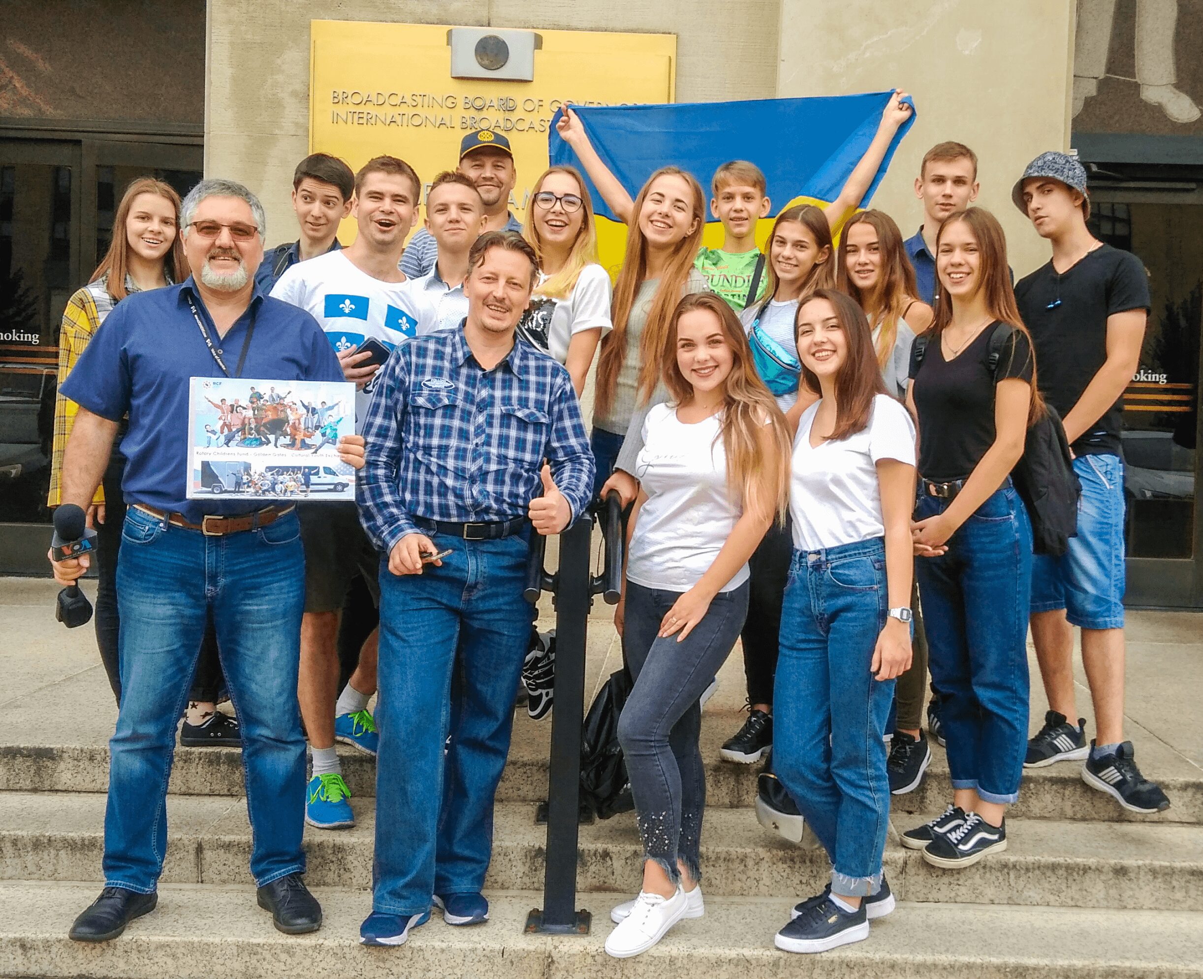 Group of young people and adults standing on steps outdoors, holding a flag and a poster, smiling.