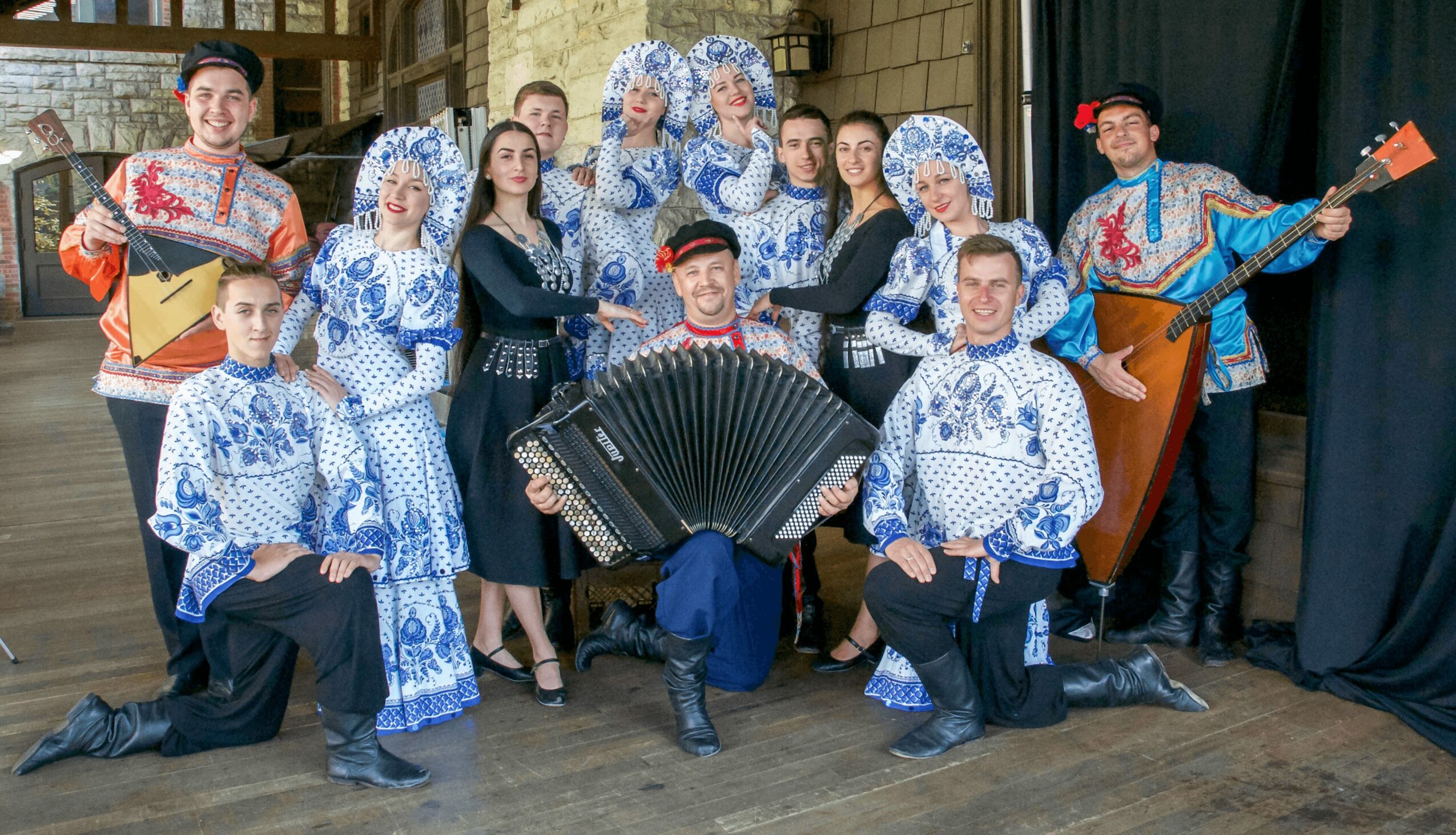 Group of people in traditional costumes, some holding musical instruments, posing indoors with a wooden background.