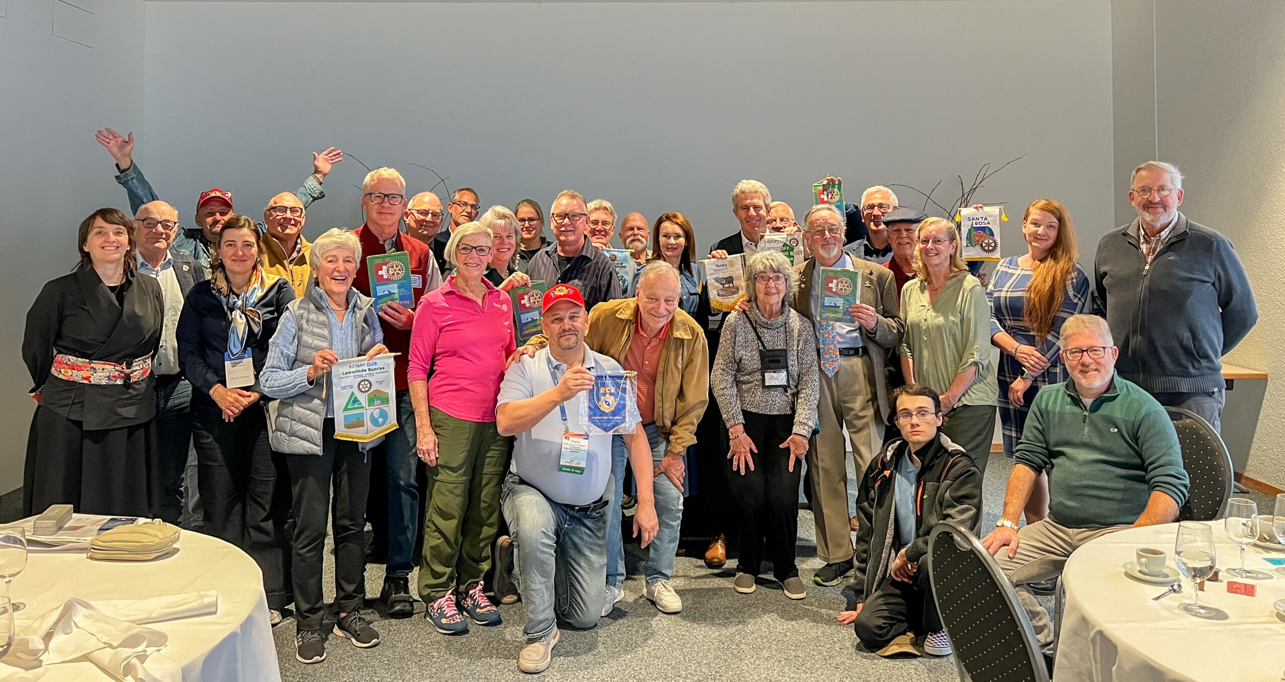 Group of people gathered in a room, some holding gift bags, smiling for a photo, with tables and chairs nearby.