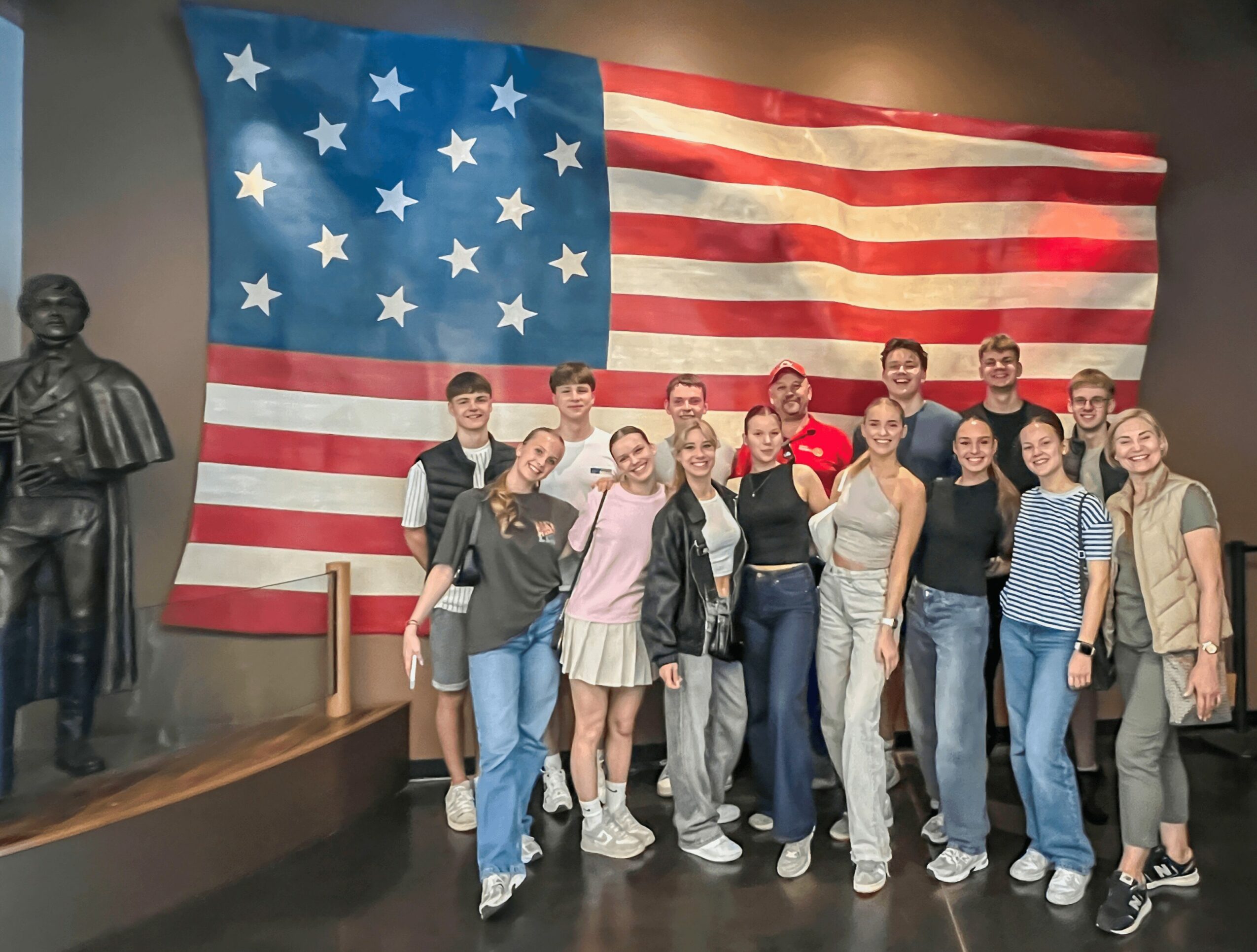 Group of people standing in front of a large American flag display, with a statue on the left side.