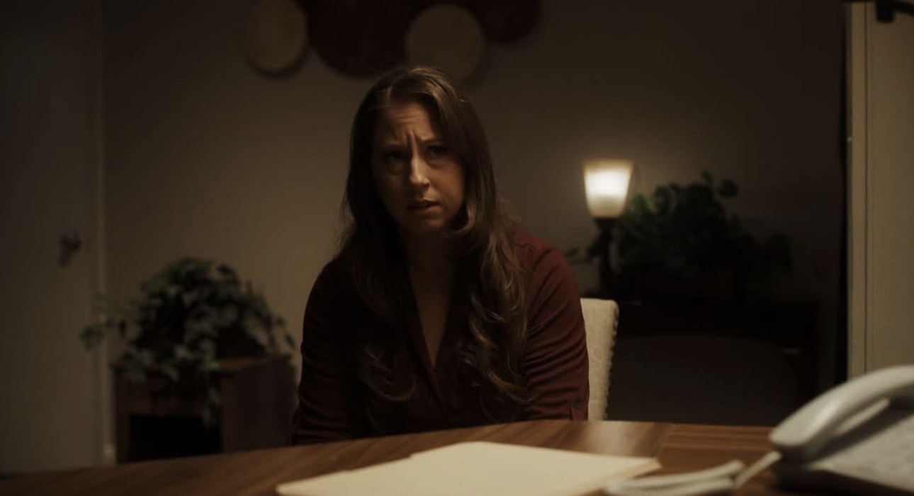 Woman with long hair sitting at a table in a dimly lit room, looking concerned, with papers and a phone on the table.