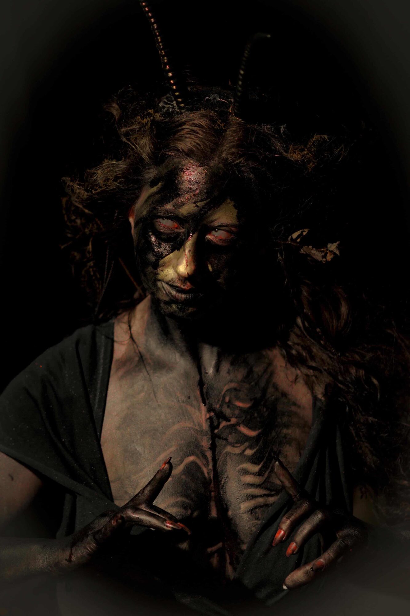 Person with face and body painted, wearing antennae, in dark lighting, with a serious expression, holding a finger up.