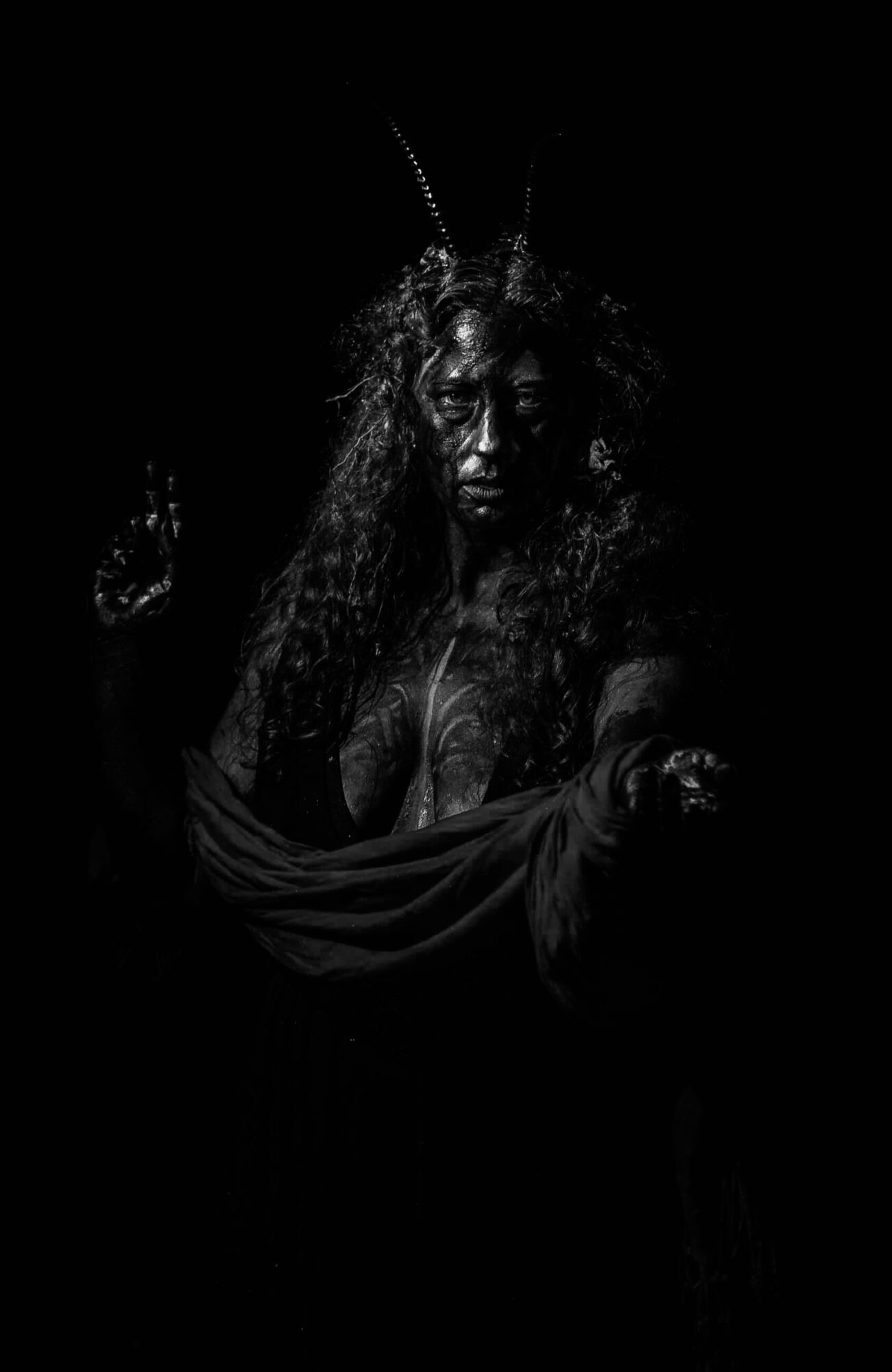 A person with long, curly hair and face paint, wearing a headpiece with feathers, in a dark setting.