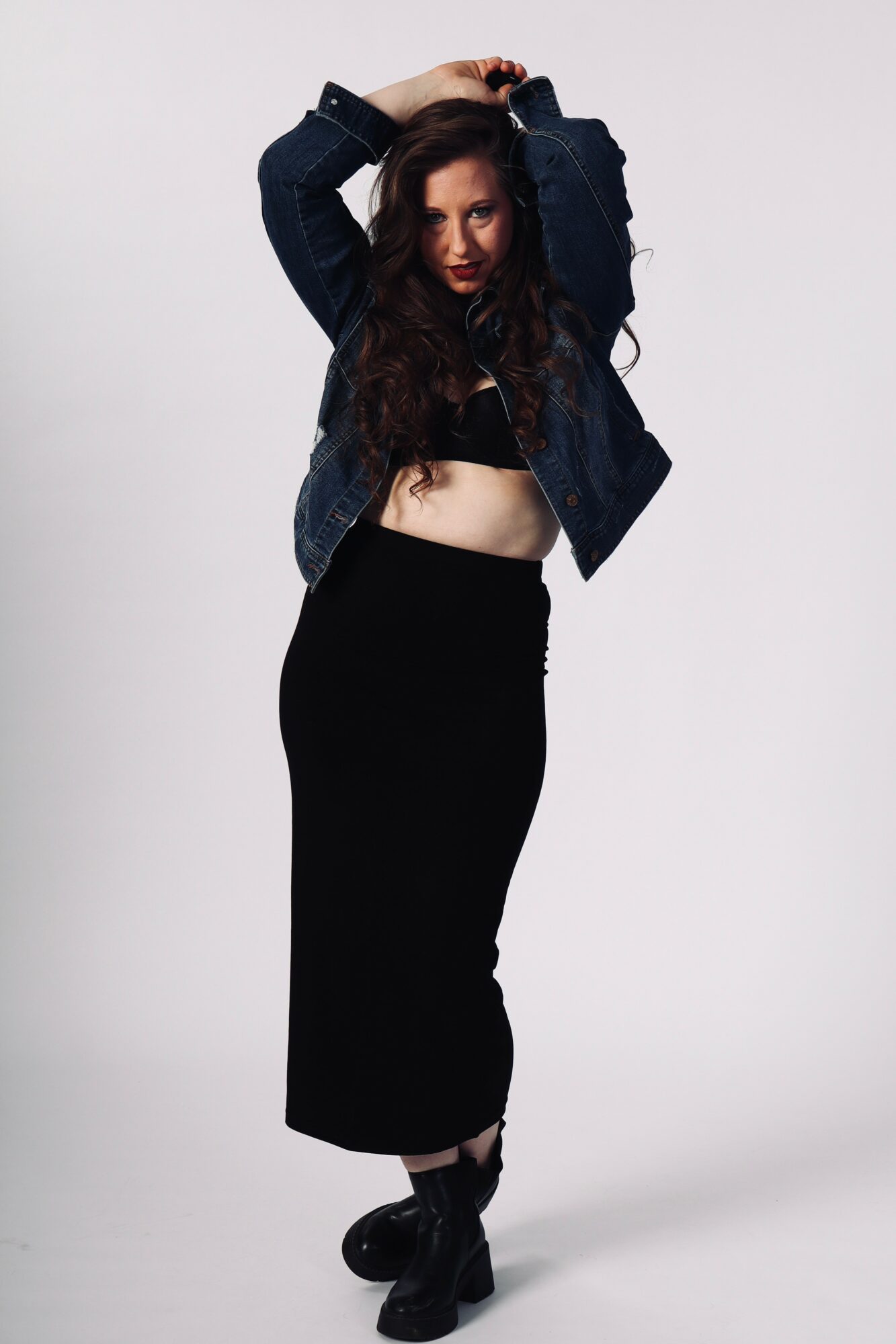 Woman with long dark hair wearing a denim jacket, black crop top, and black skirt, posing with arms raised above head.