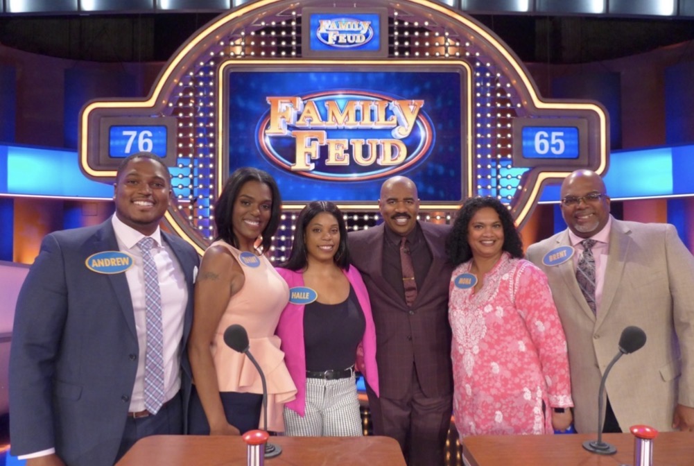 Six people stand behind a game show podium with a 'Family Feud' sign in the background.