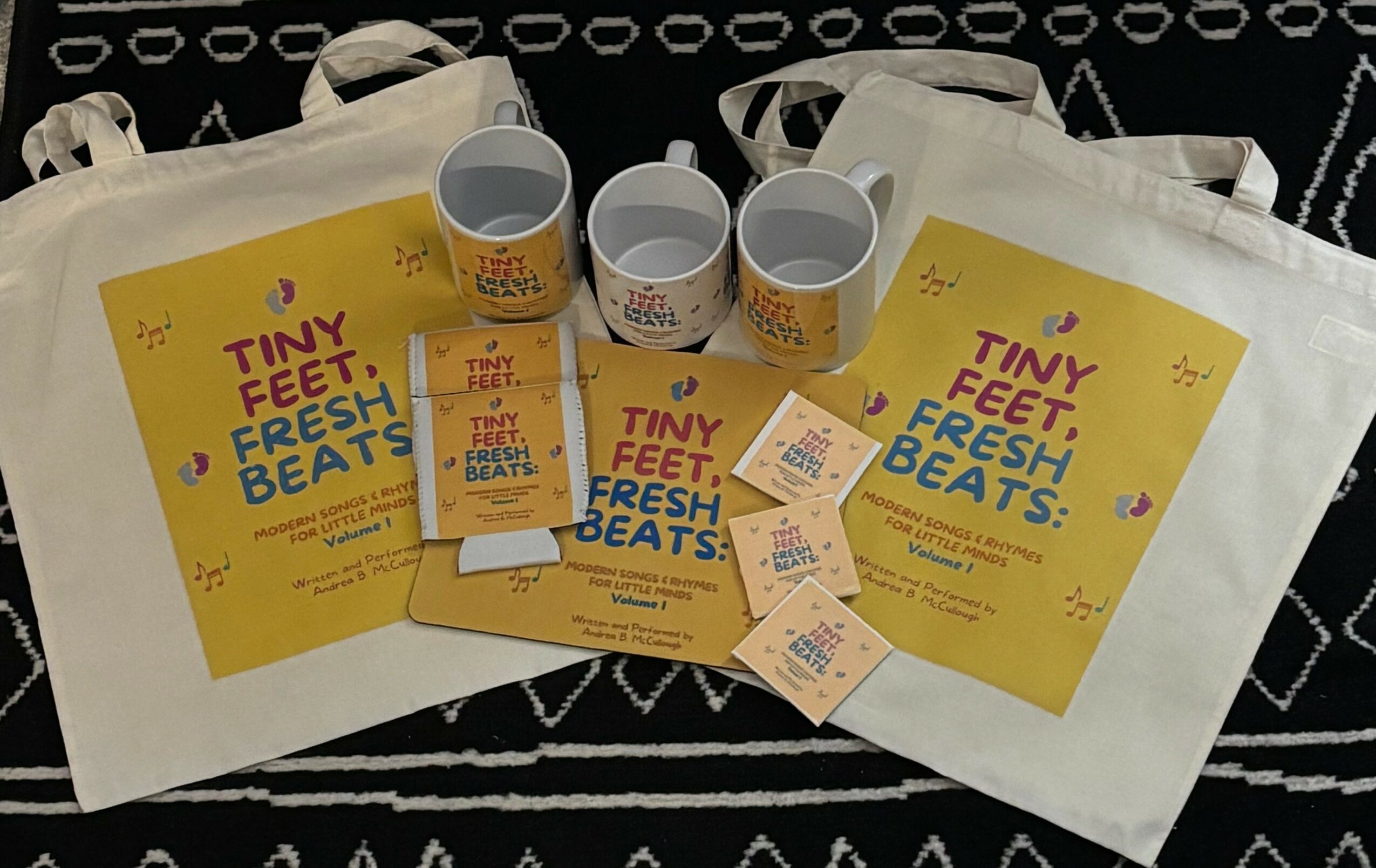 Two white tote bags, two cups, and two flyers with yellow backgrounds and colorful text, on a black surface.