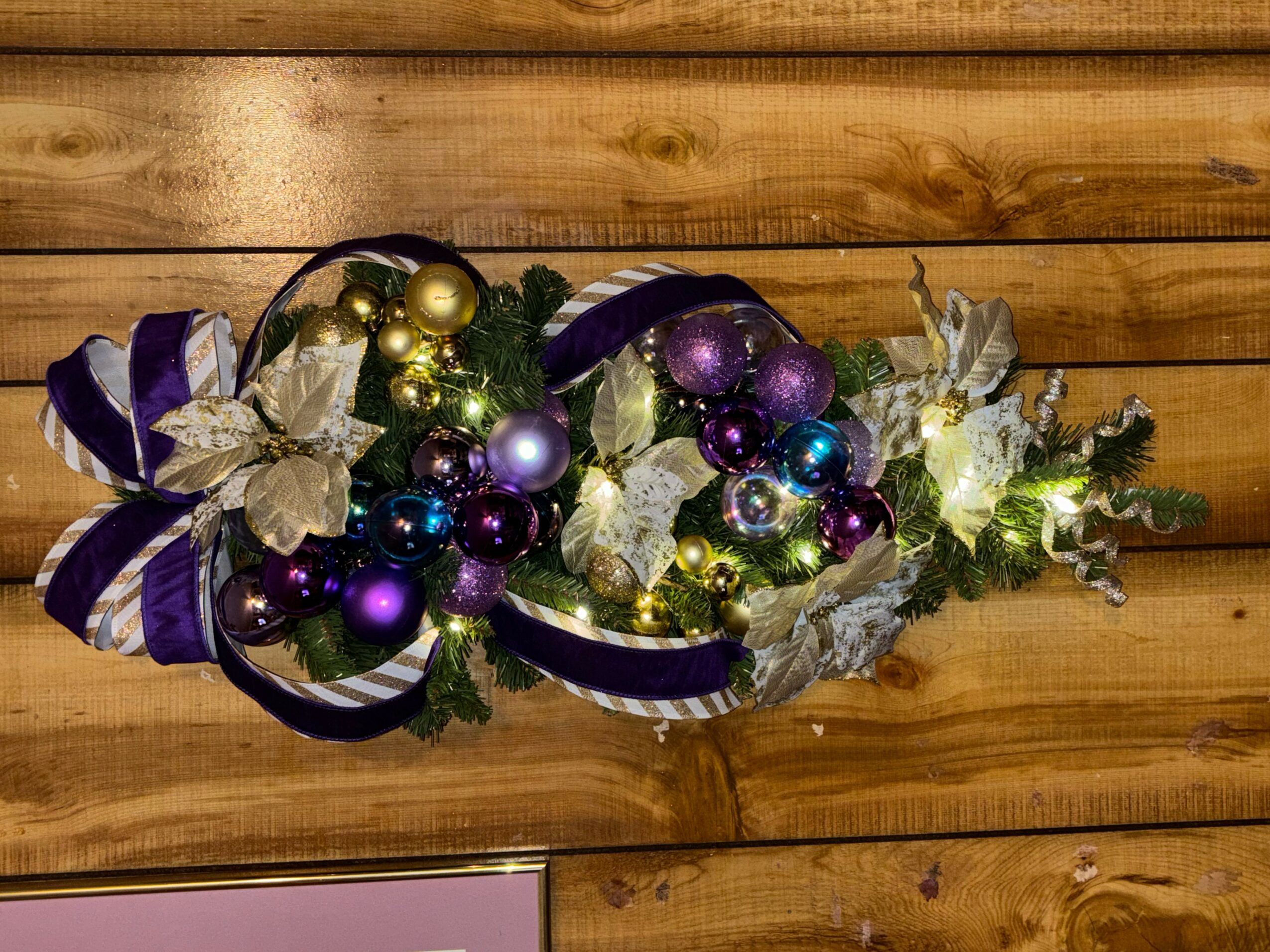 Decorative arrangement with purple, gold, and silver ornaments, ribbons, and foliage on a wooden surface.