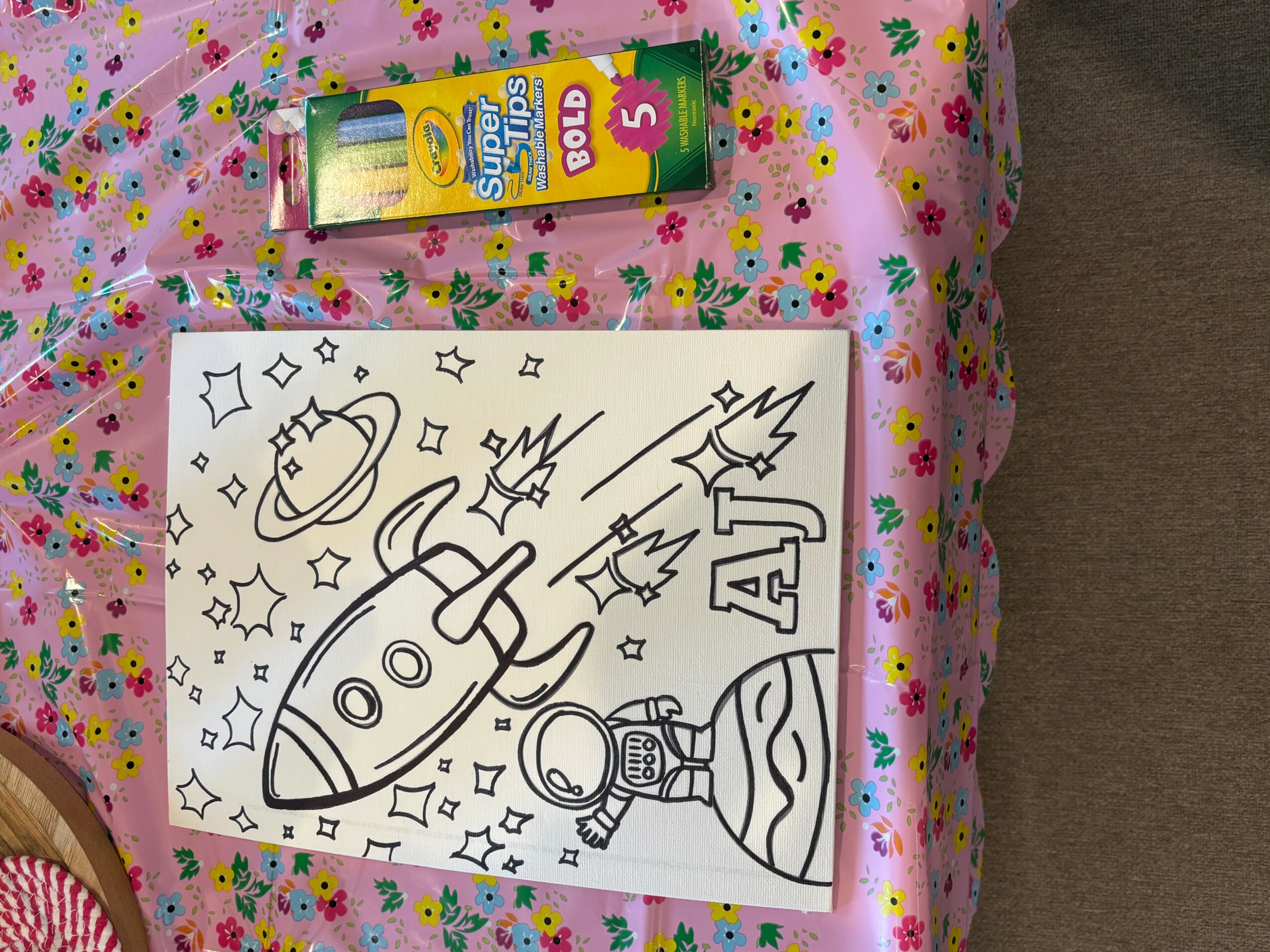 Coloring page with a rocket, astronaut, and planets, surrounded by stars and space objects, on a pink floral tablecloth.