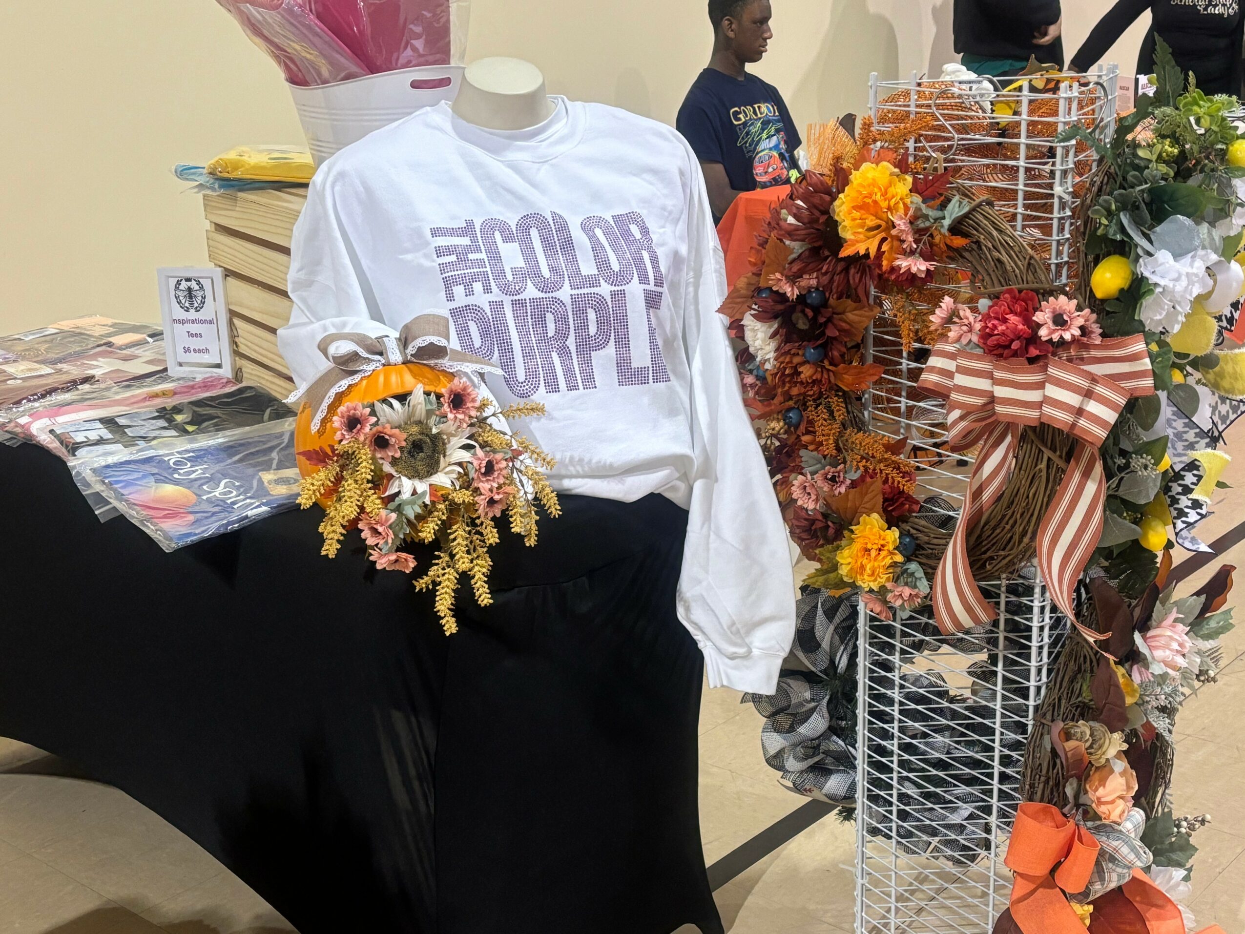 Mannequin wearing a white sweatshirt with purple text, surrounded by floral arrangements and display items, indoors.