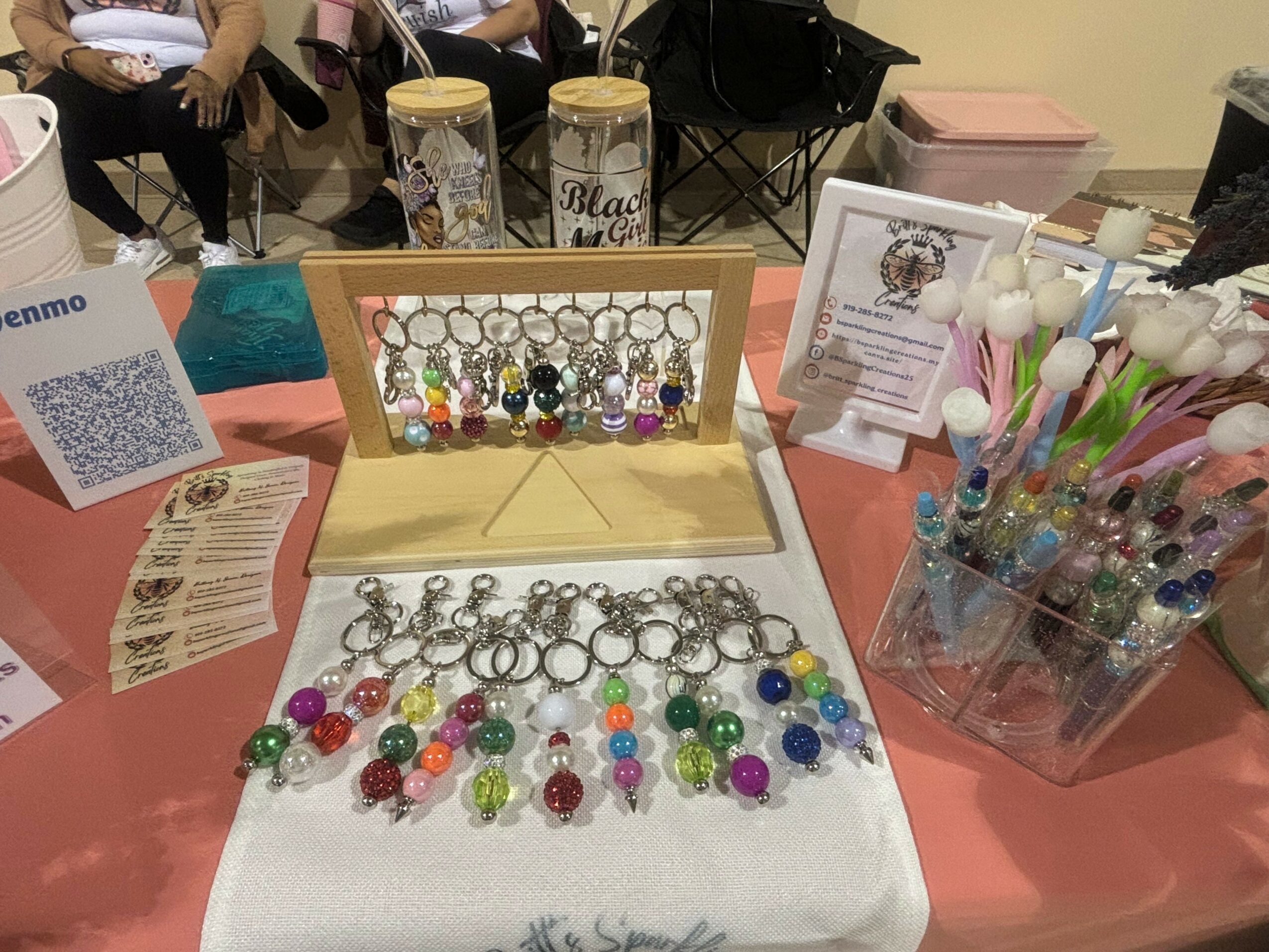Display of colorful beaded jewelry on a table with a white cloth, including rings and earrings, with other craft items around.