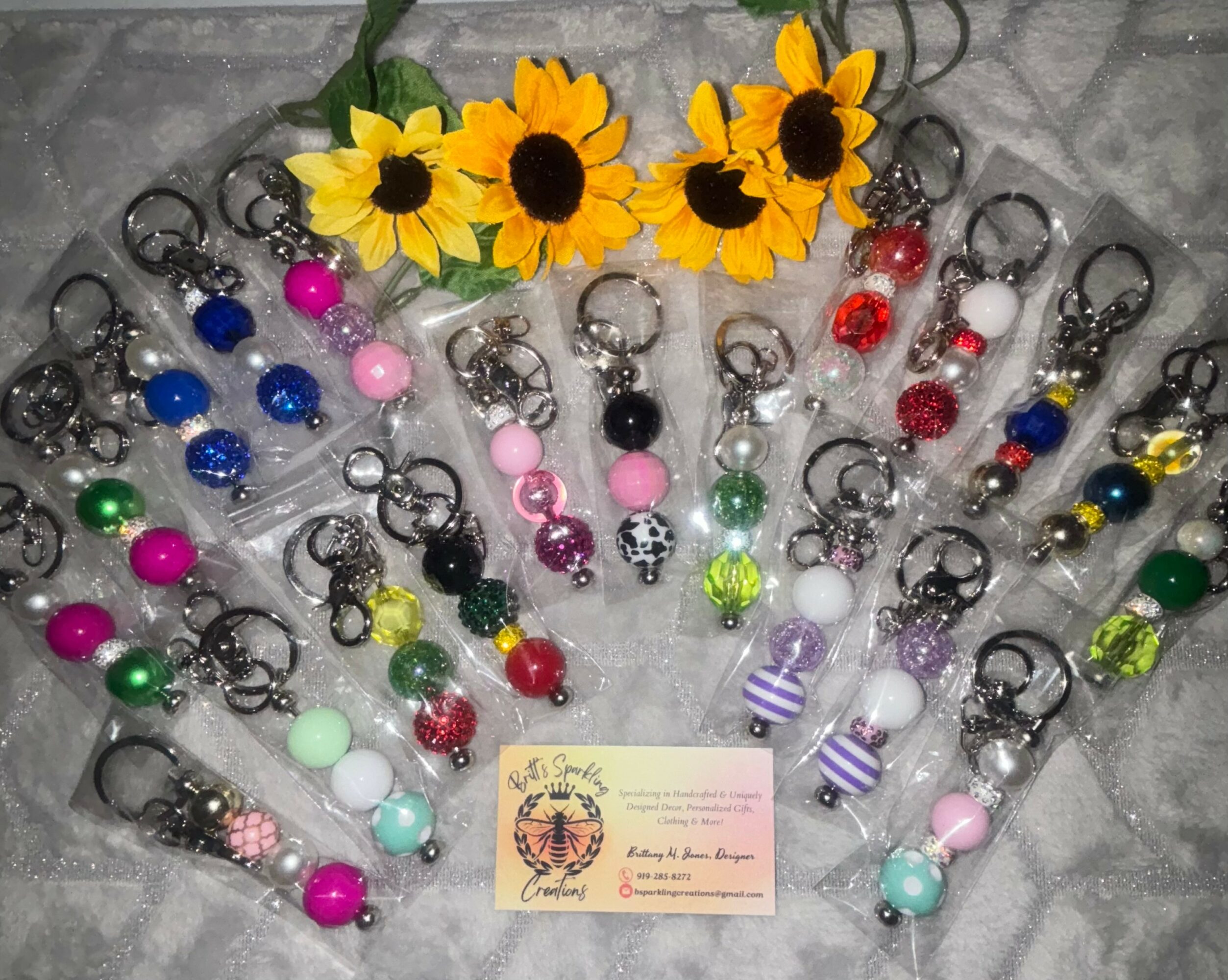 Collection of colorful keychains with beads and charms, yellow flowers, and a business card on a gray surface.