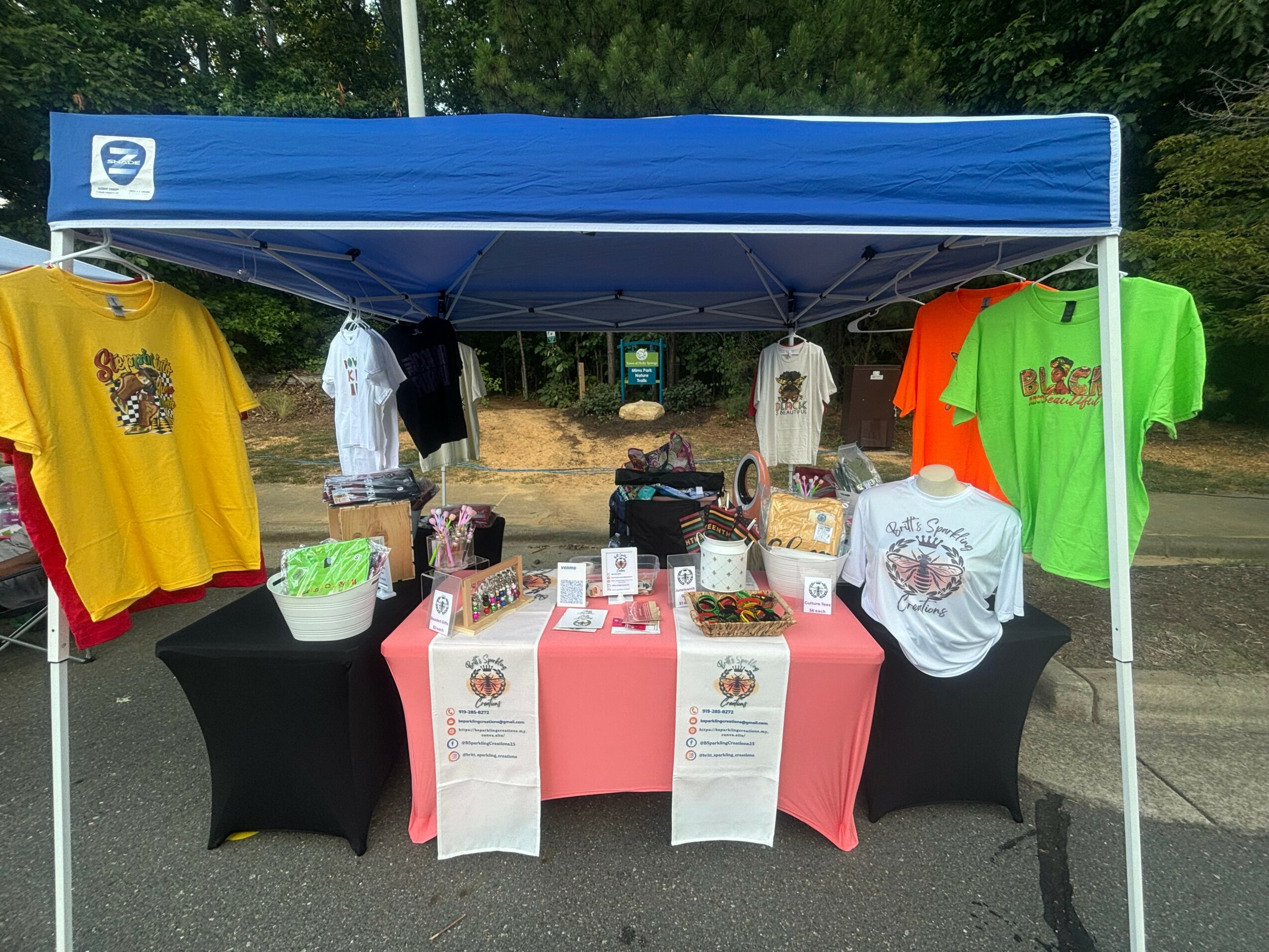Outdoor booth with T-shirts on display, table with promotional materials, under a blue canopy, trees in background.