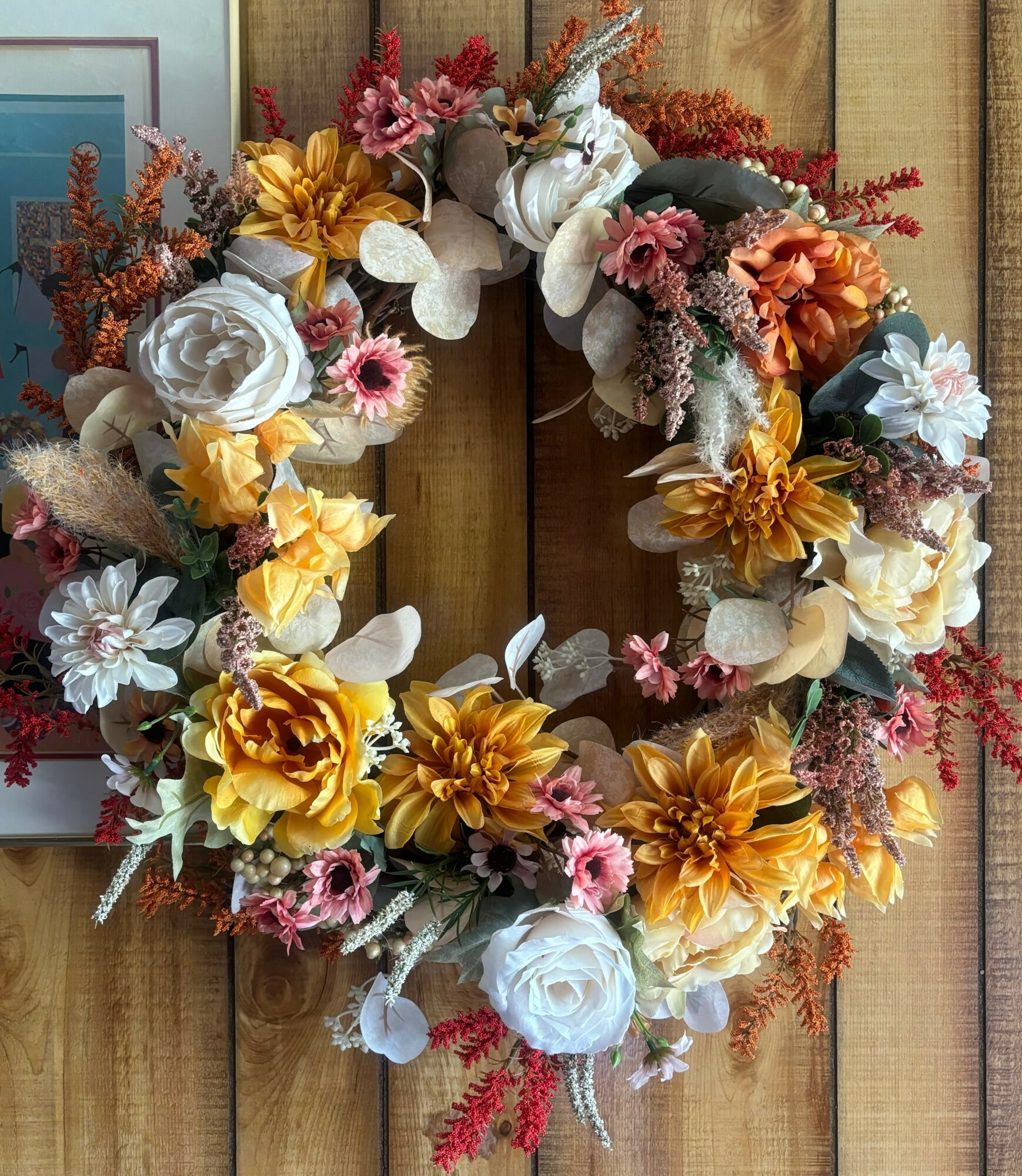 Decorative floral wreath with white, yellow, pink, and orange flowers hanging on a wooden surface.