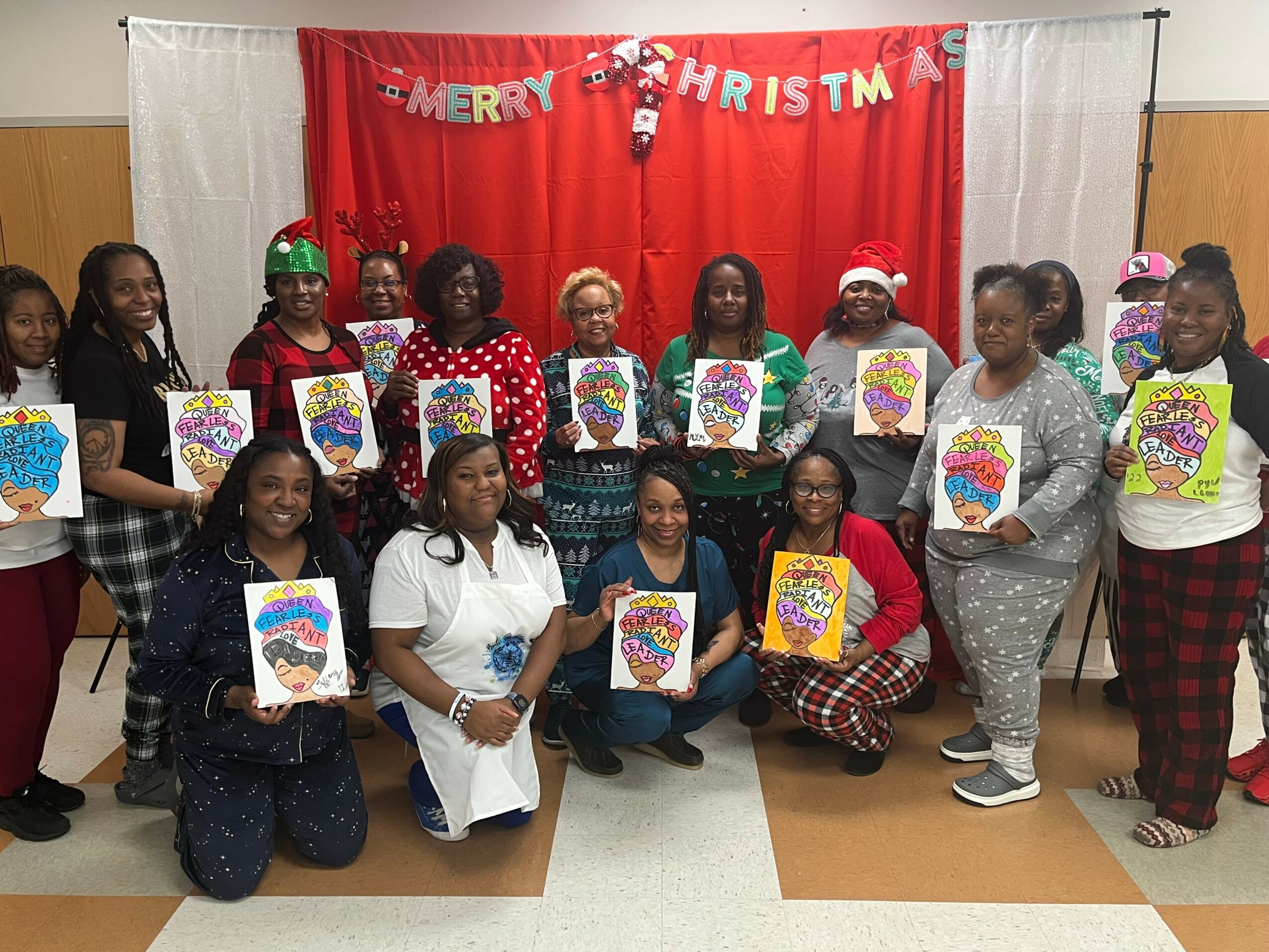 Group of women and girls holding Christmas-themed coloring pages in front of a red and white holiday backdrop with 'Merry Christmas' banner.
