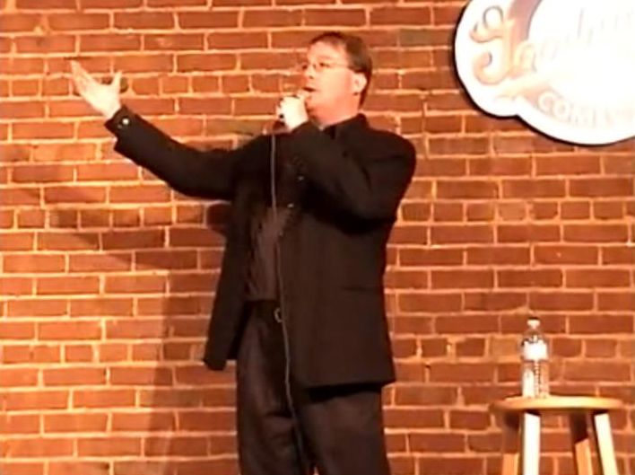 Person performing stand-up comedy on stage with brick wall background, holding microphone, gesturing with one hand, water bottle on stool