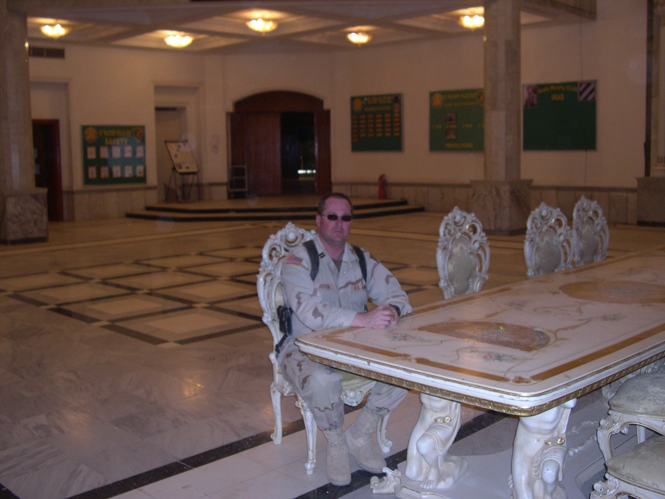 Person sitting at a large ornate table in a spacious room with high ceilings and green wall displays.