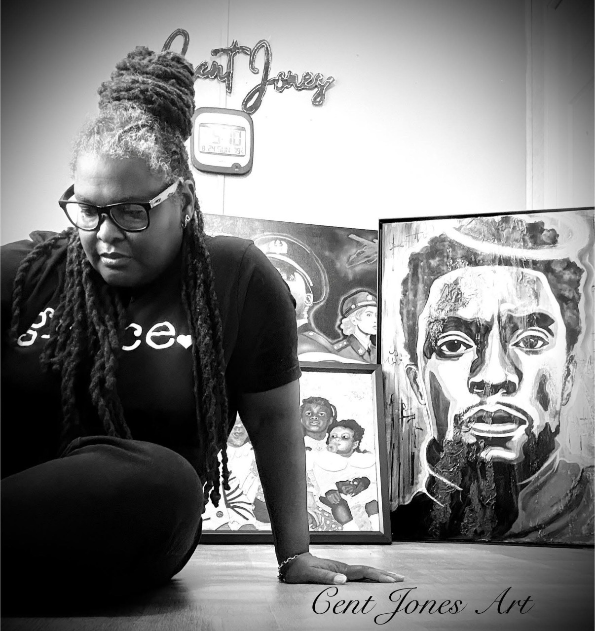 Person with glasses and dreadlocks sitting on the floor, art displayed behind, black and white photo.