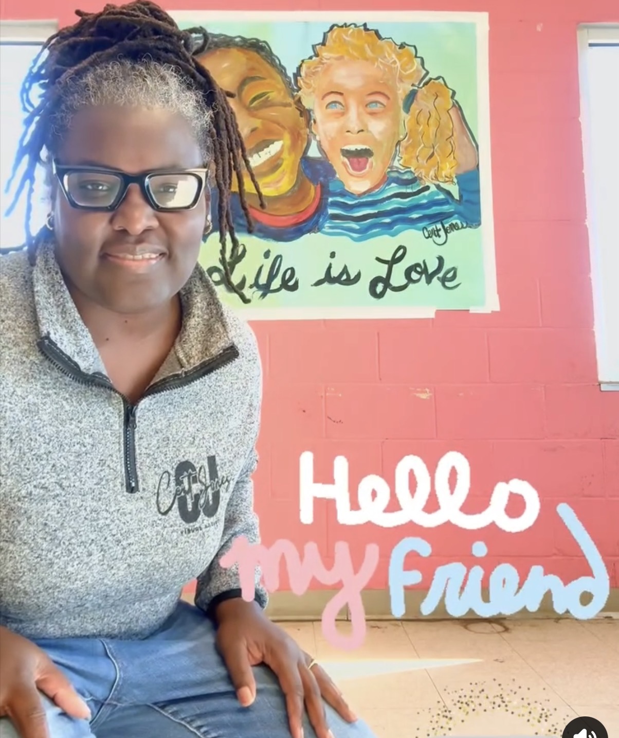 Woman with glasses and dreadlocks standing in front of a colorful mural, with text saying 'Hello my Friend'.