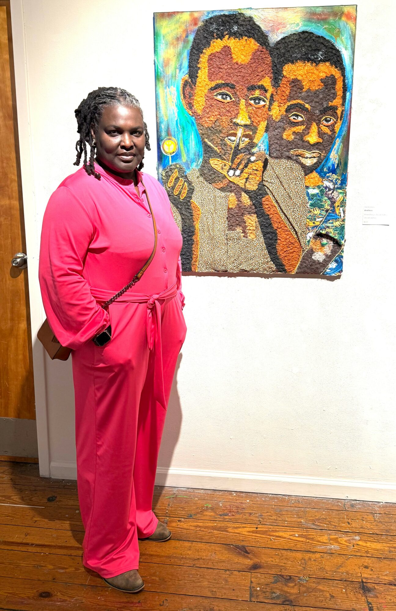 Woman in pink jumpsuit standing next to colorful portrait of two men on wall, wooden floor, white wall background.