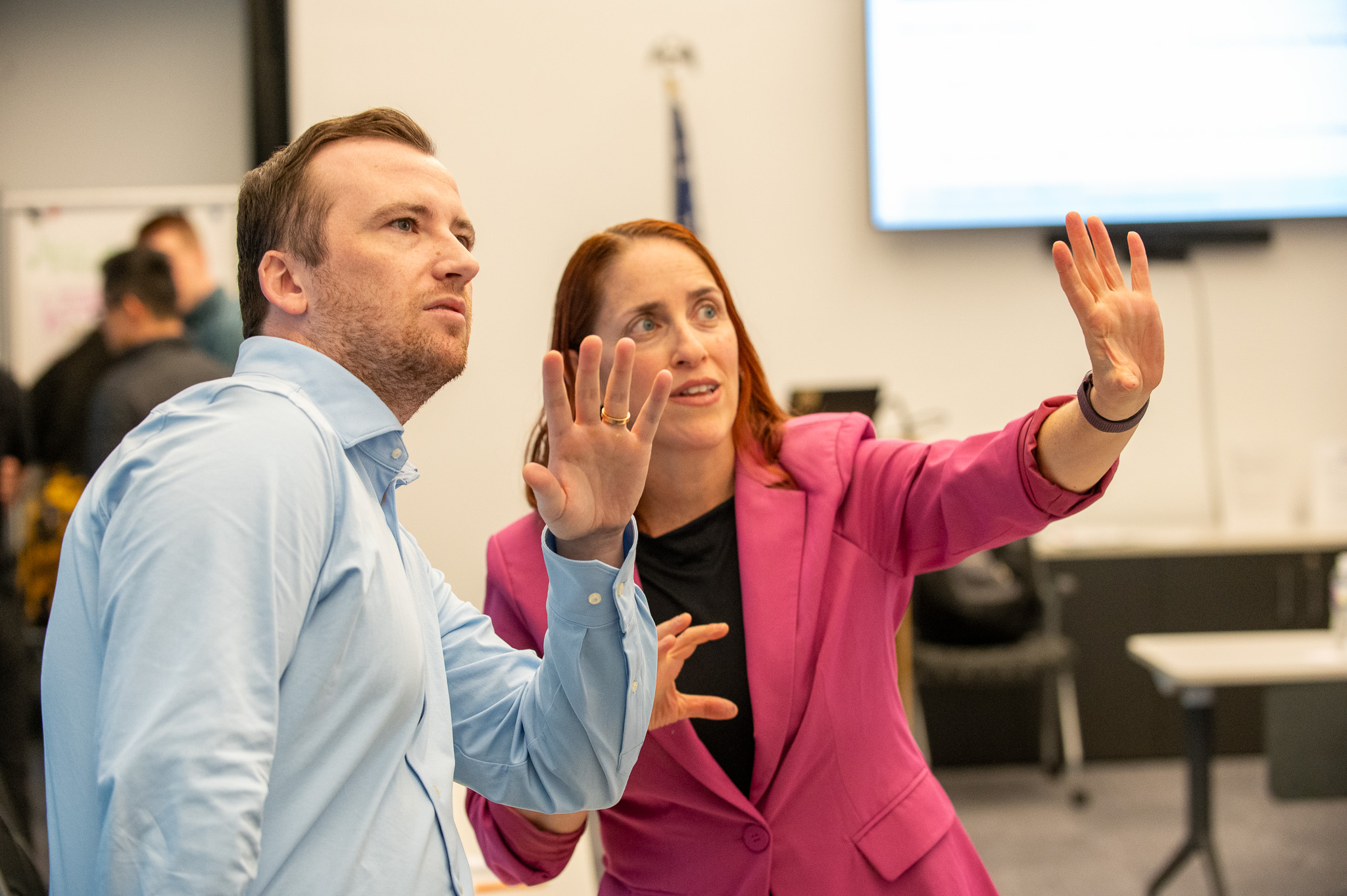 Two people in a meeting room, one woman gesturing with her hands, a man listening attentively, blurred background with a screen.