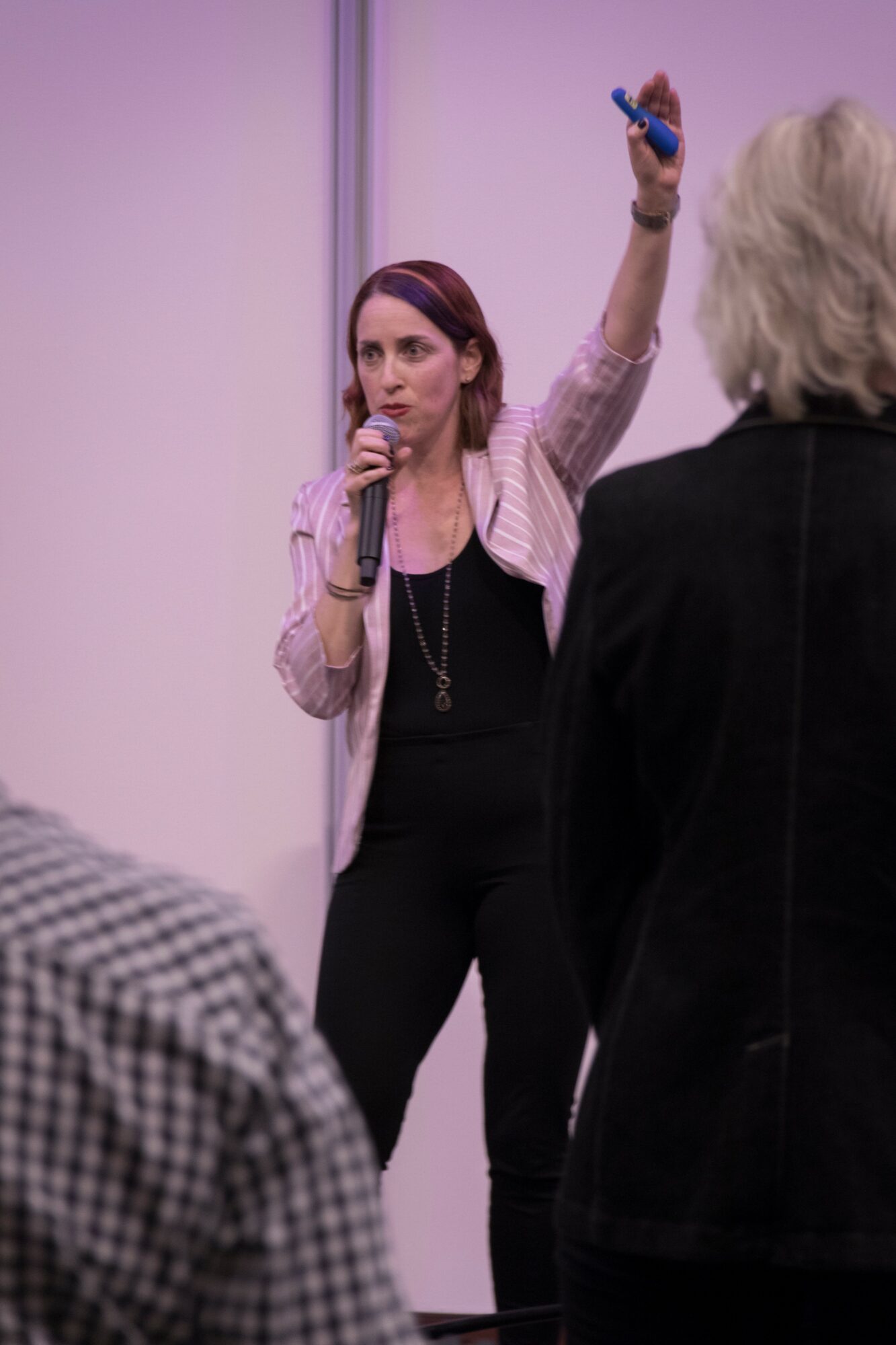 Woman speaking into microphone, raising a hand with a marker, in front of a group, in a presentation setting.