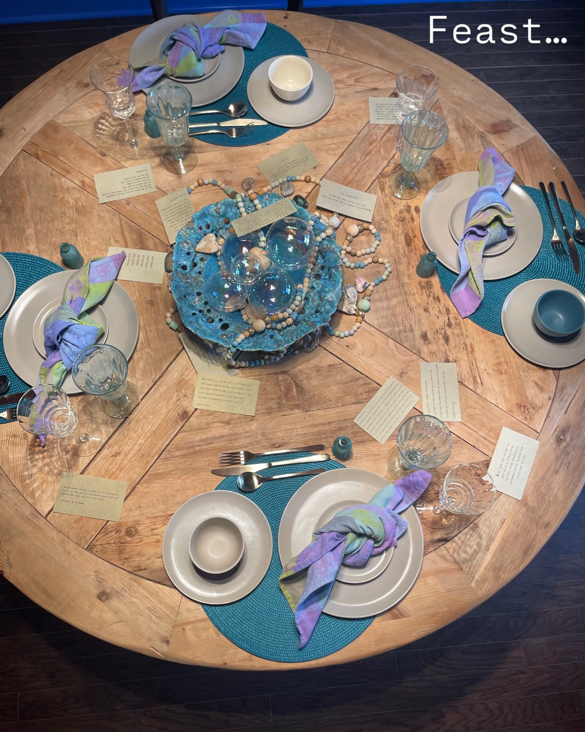 Round wooden table set for a feast with plates, glasses, cutlery, and decorated napkins, with a blue cake in the center.