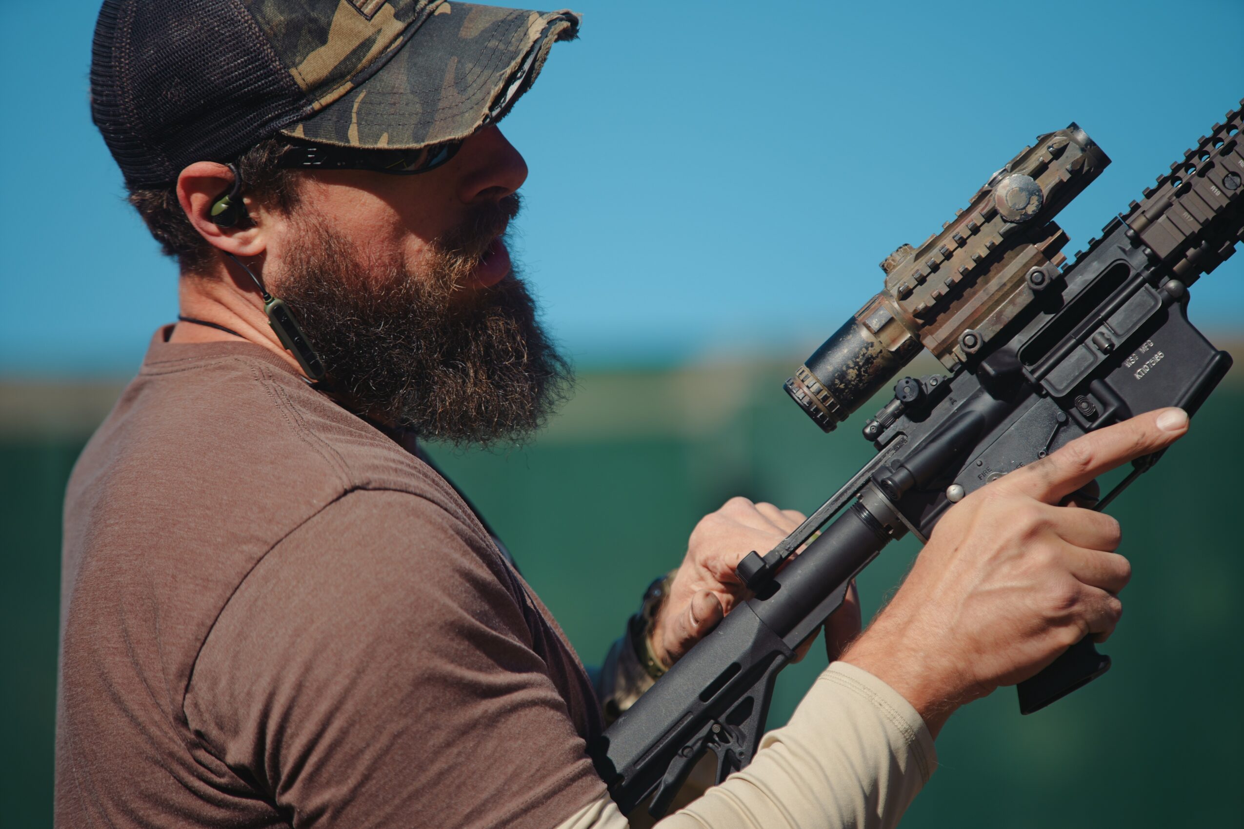 Man with a beard, wearing a cap, holding a rifle with a scope, outdoors against a blue sky background.