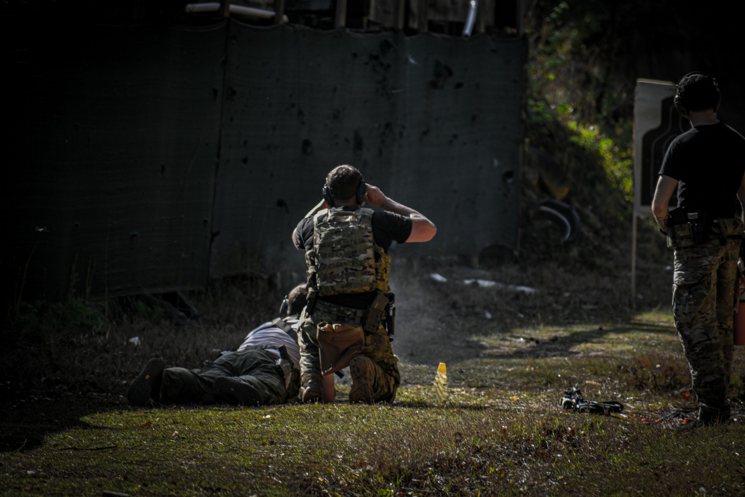 Two armed individuals detain a person lying on the ground outdoors near a dark wall, with another person standing nearby.