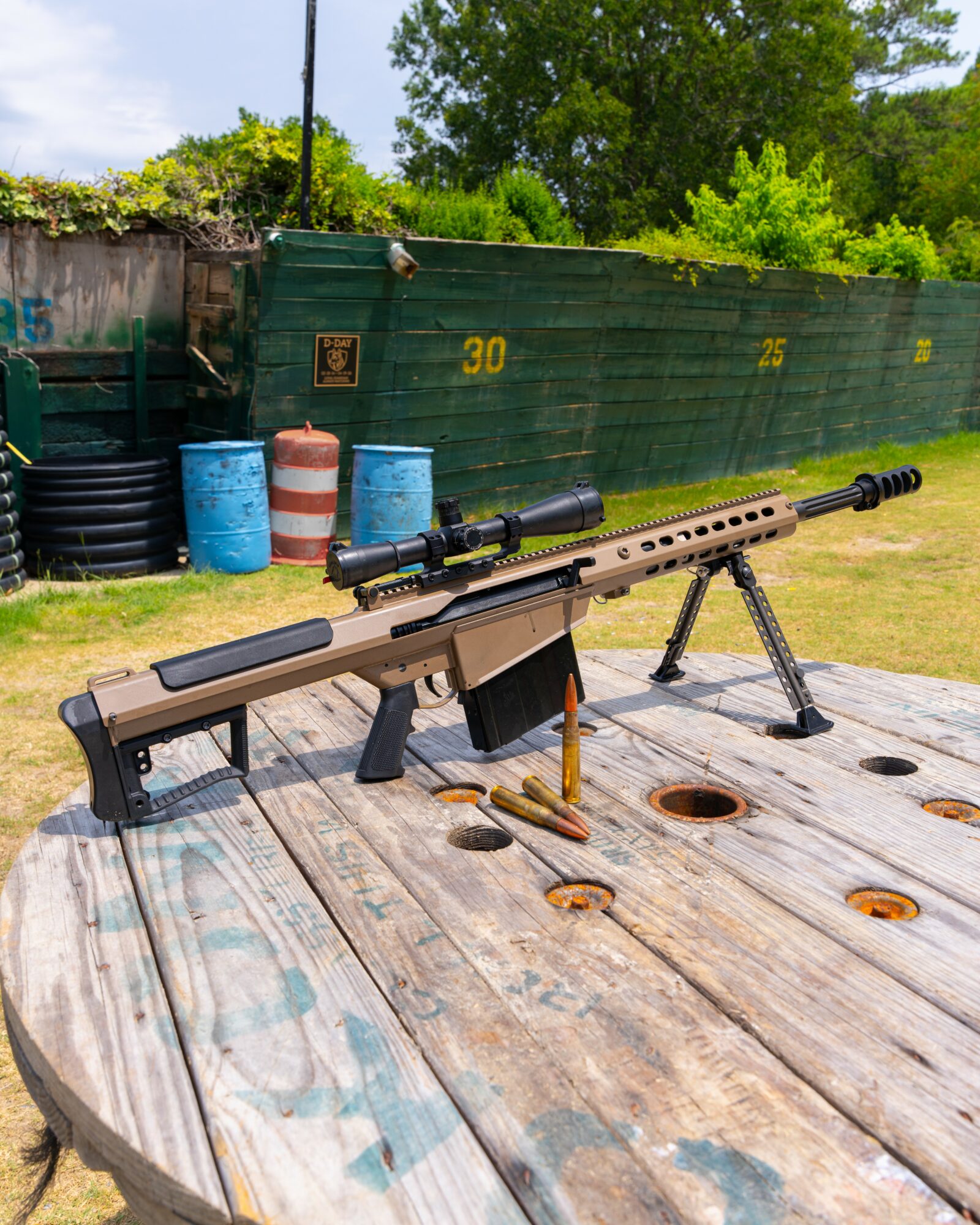 Sniper rifle with scope and bipod on wooden surface outdoors, background includes green fence, barrels, and trees.