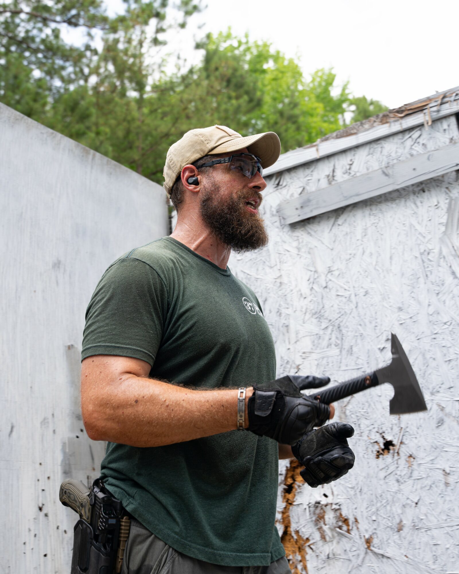 Man with beard, sunglasses, and cap holding a hammer outdoors, standing near a white wall with trees in background.