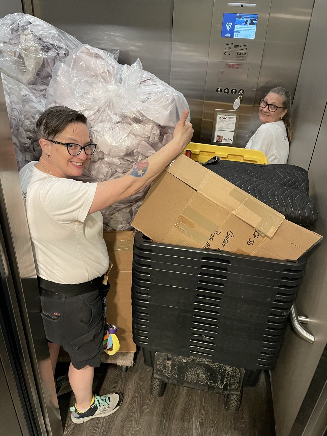 Two women in an elevator with large bags and boxes, smiling and handling items. One woman is near the elevator wall, the other in the background.