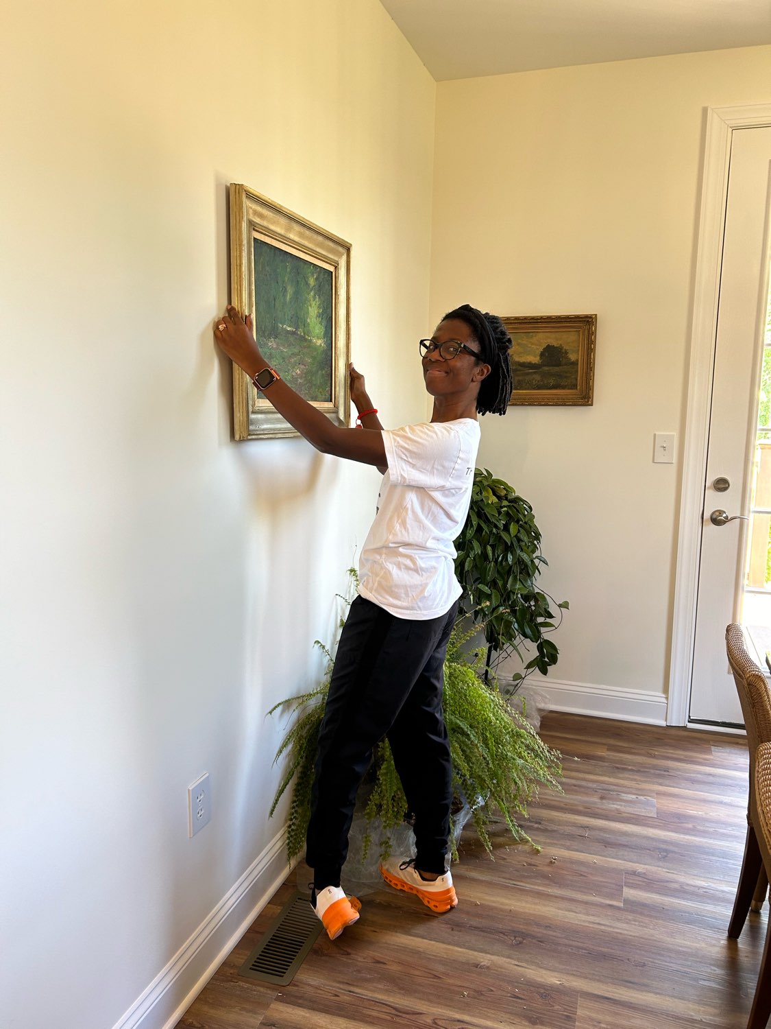 Woman hanging a framed painting on a wall in a room with hardwood floor and potted plants.