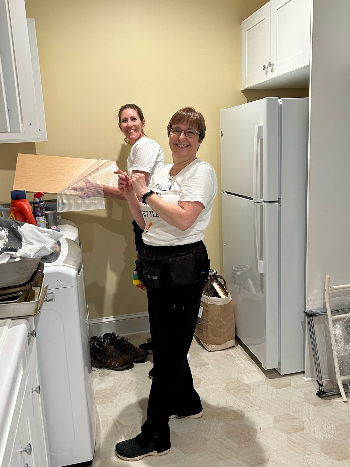 Two women in a kitchen, one smiling and clapping, the other preparing food at the counter, with appliances and cabinets around.