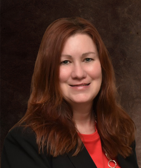 Portrait of a woman with shoulder-length reddish-brown hair, smiling, wearing a black jacket and red top, against a dark background.