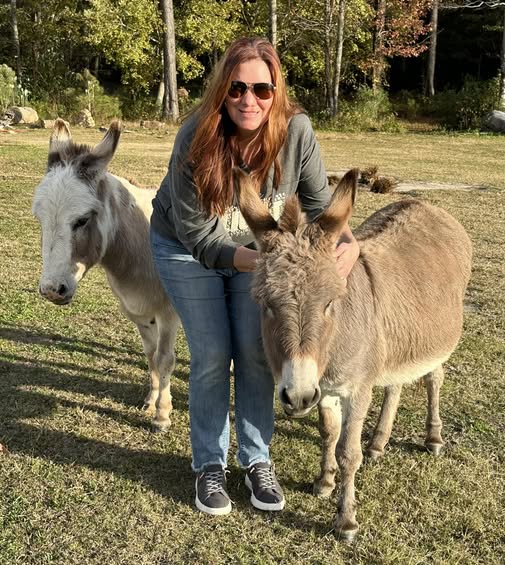 Woman with sunglasses crouching next to two donkeys in a grassy field, trees in the background.