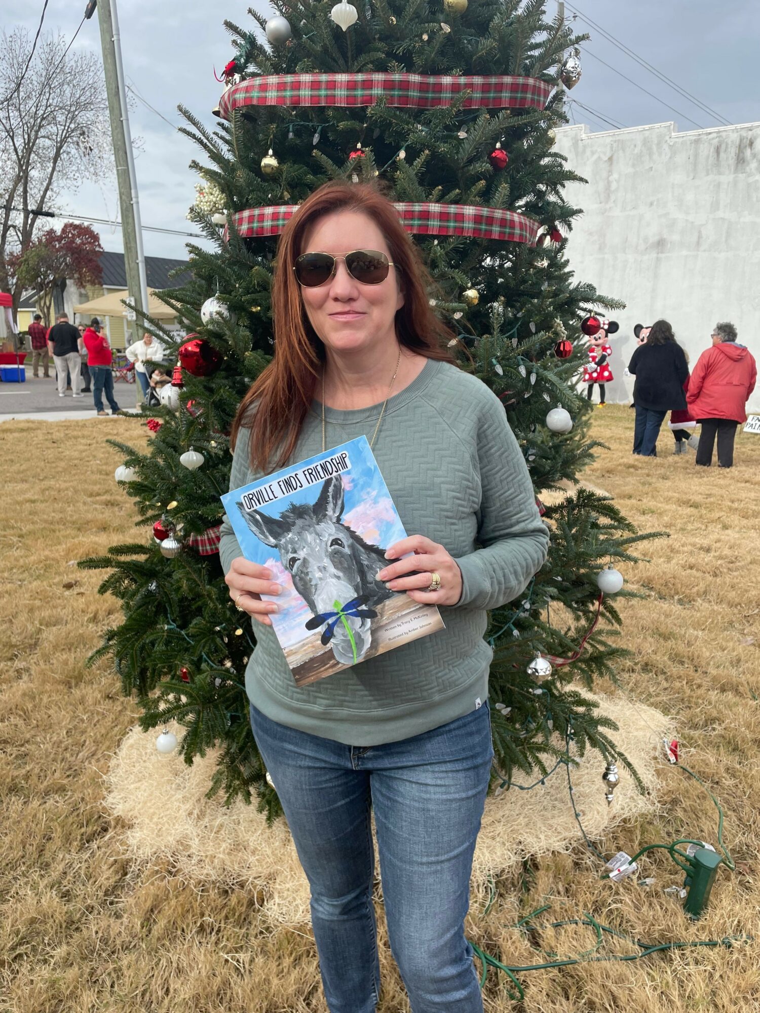 Woman with sunglasses holding a magazine in front of a decorated Christmas tree outdoors, with people in the background.