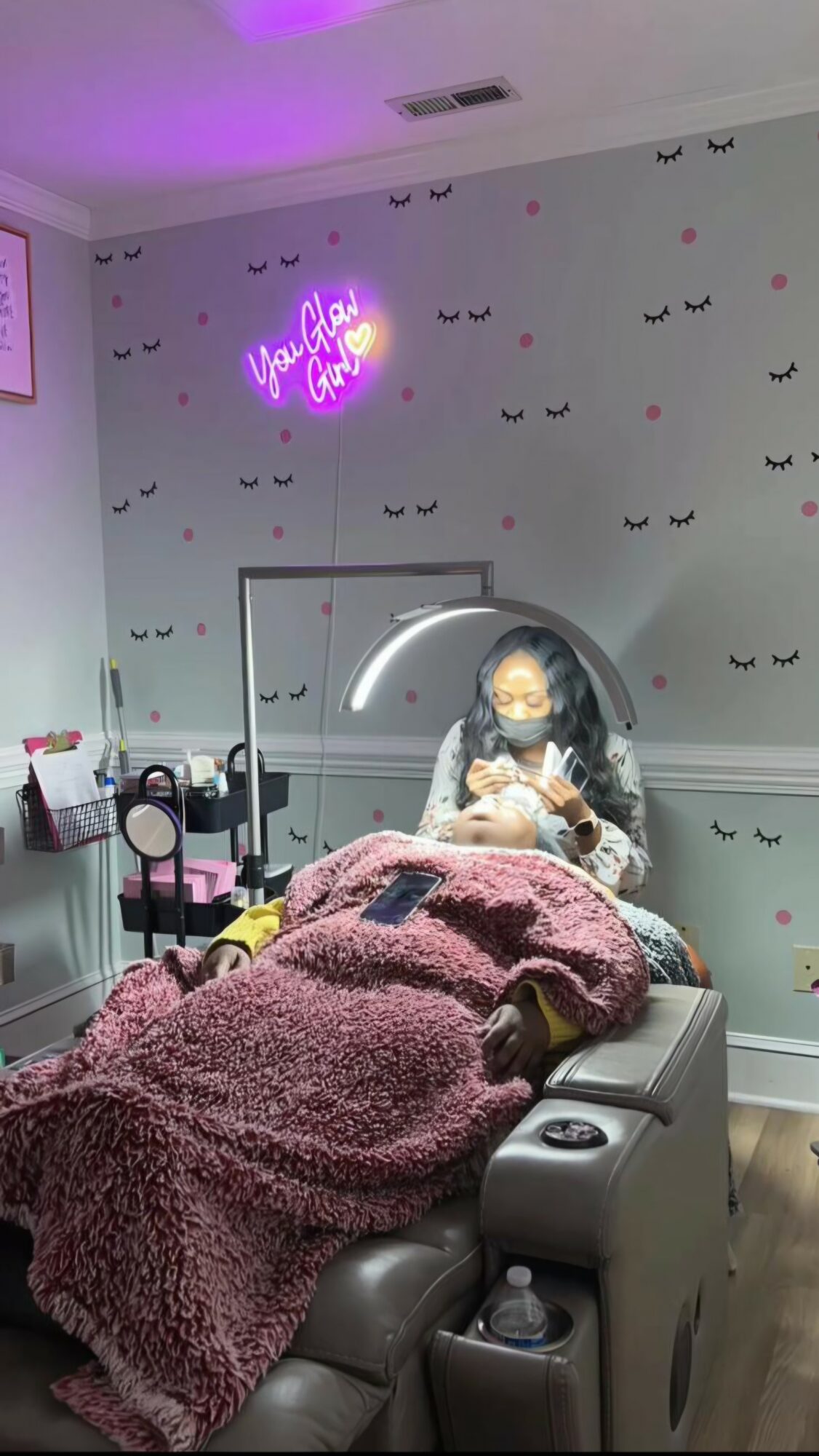 Person lying on a bed in a room with wall decorations and a neon sign, taking a mirror selfie.