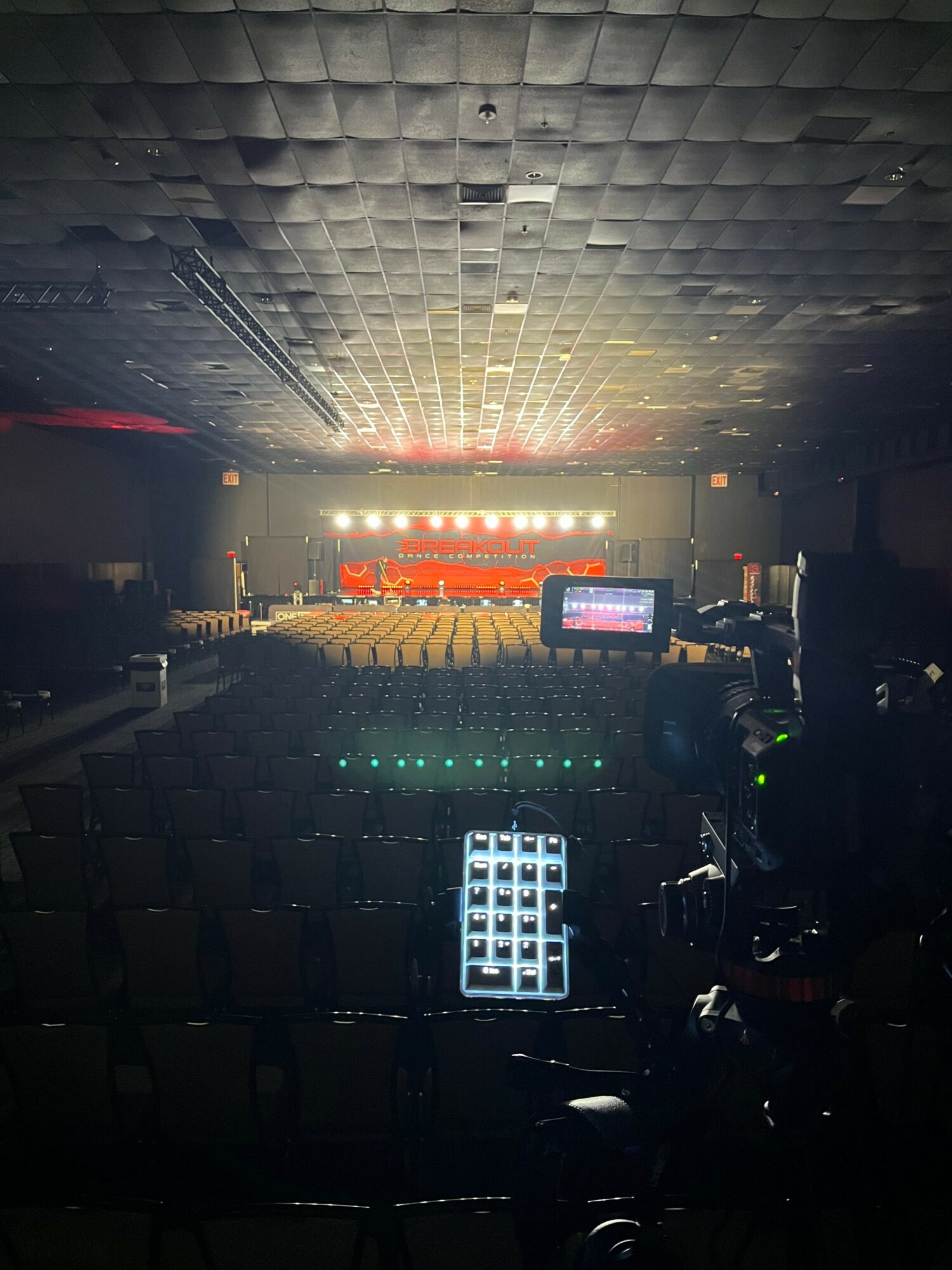 Empty auditorium with stage lights and seating, view from behind the control booth, equipment visible in foreground.