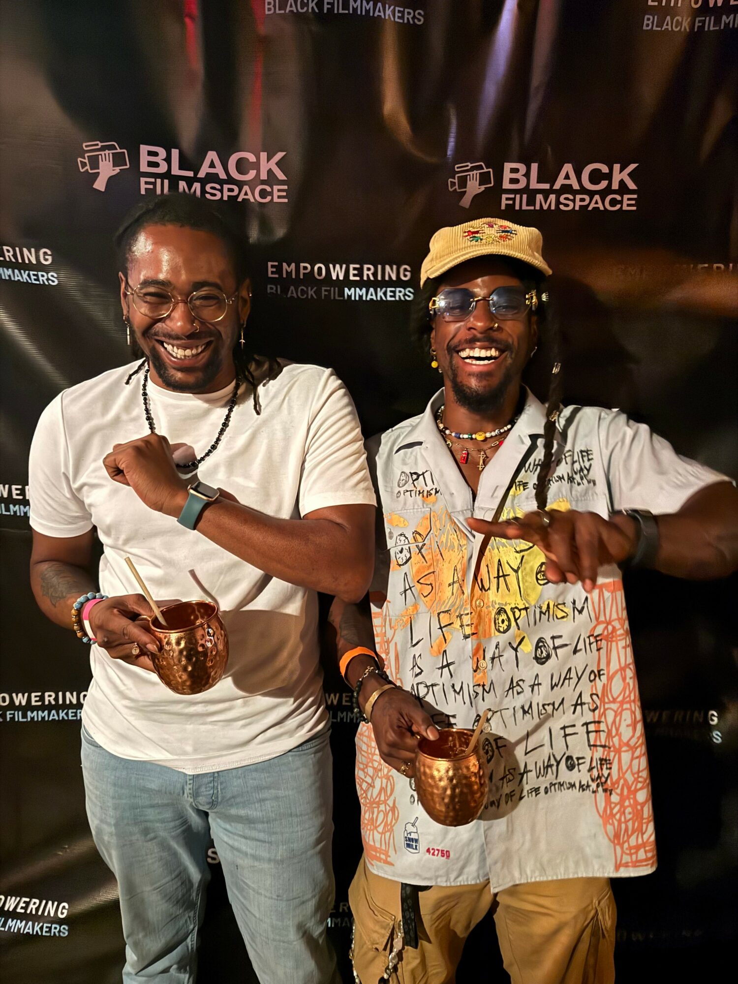 Two smiling men holding awards and glasses, standing in front of a black backdrop with logos and text.