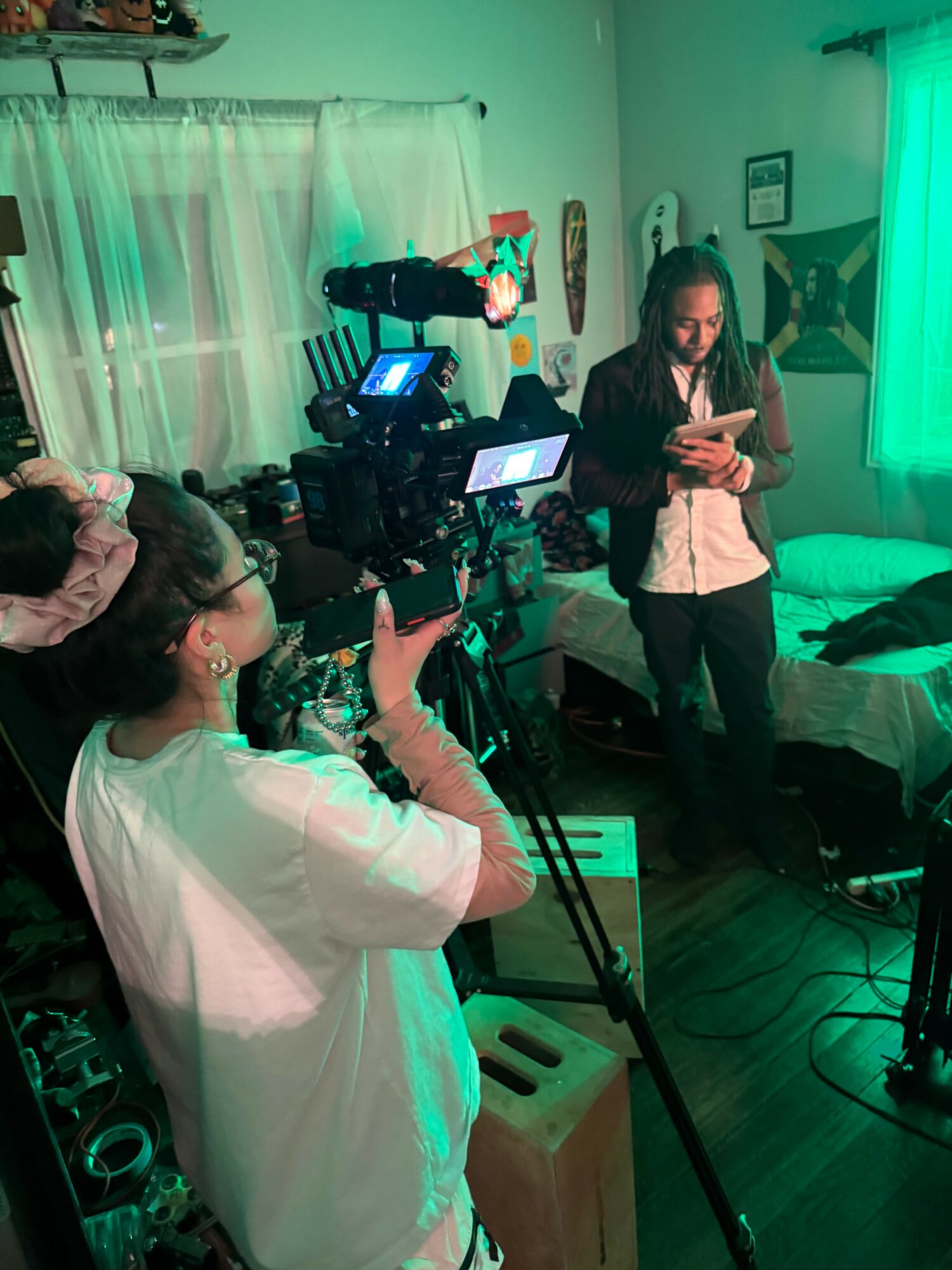 Person operating camera equipment in a room with a woman standing and using a phone, green curtains, and a couch.