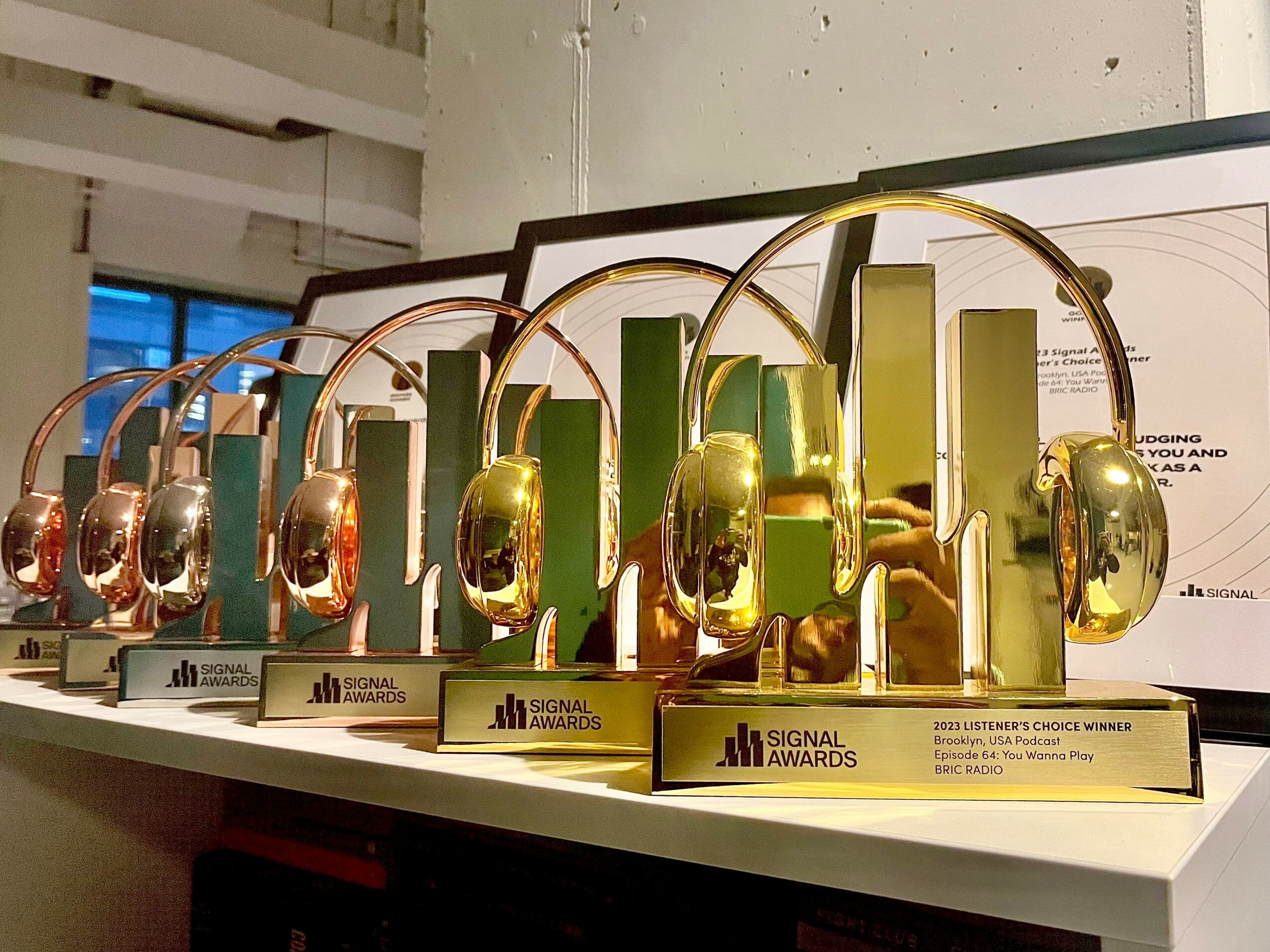 Multiple gold and bronze headphones awards displayed on a white surface, with a whiteboard and window in the background.