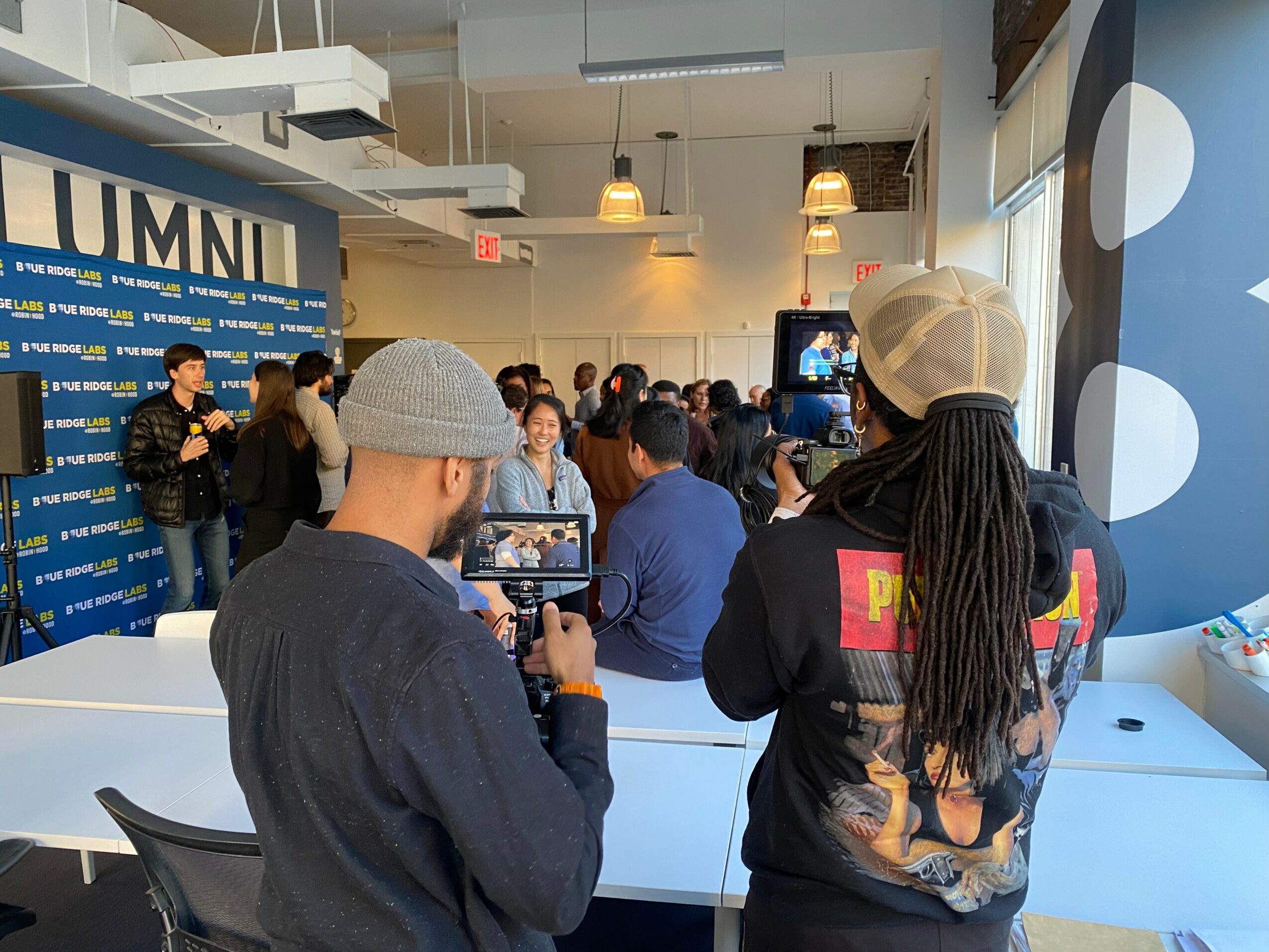 People attending an indoor event, some taking photos or videos, with a blue backdrop and a speaker at the front.
