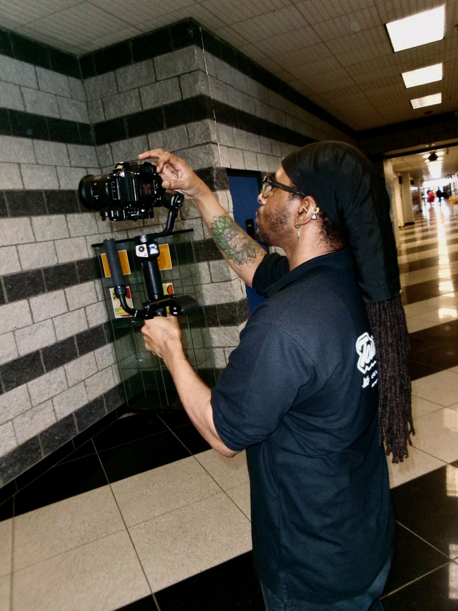 Person with long hair and glasses operating a camera mounted on a stabilizer in a hallway with black and white tiled walls.