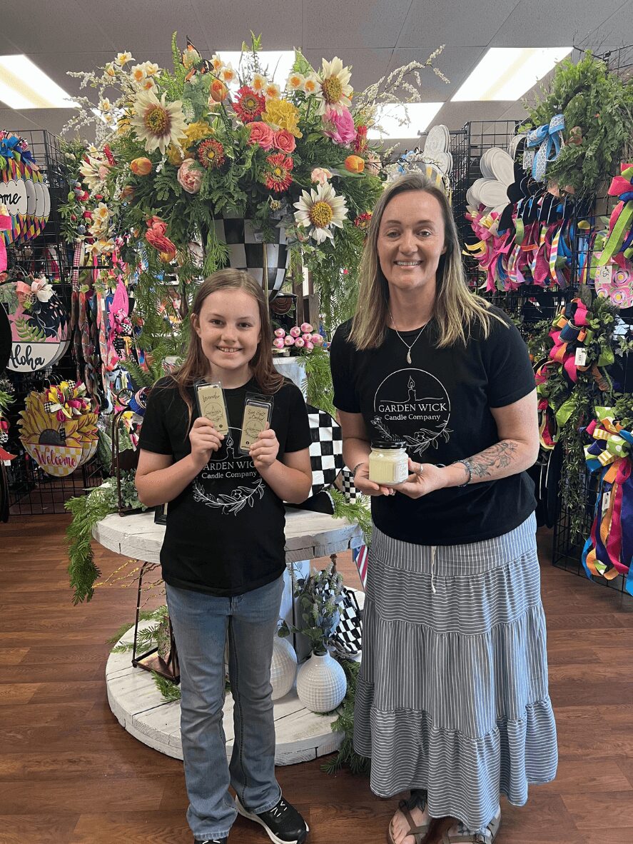 Two females, a girl and a woman, stand in a store with colorful flowers and accessories, smiling and holding small items.