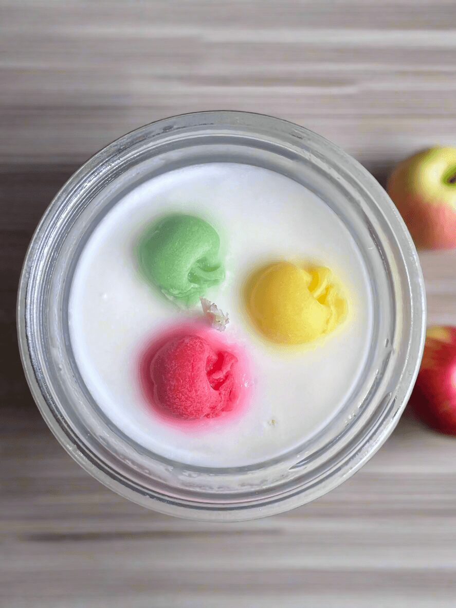 Top view of a glass jar with colorful candies in white liquid, with apples in the background.