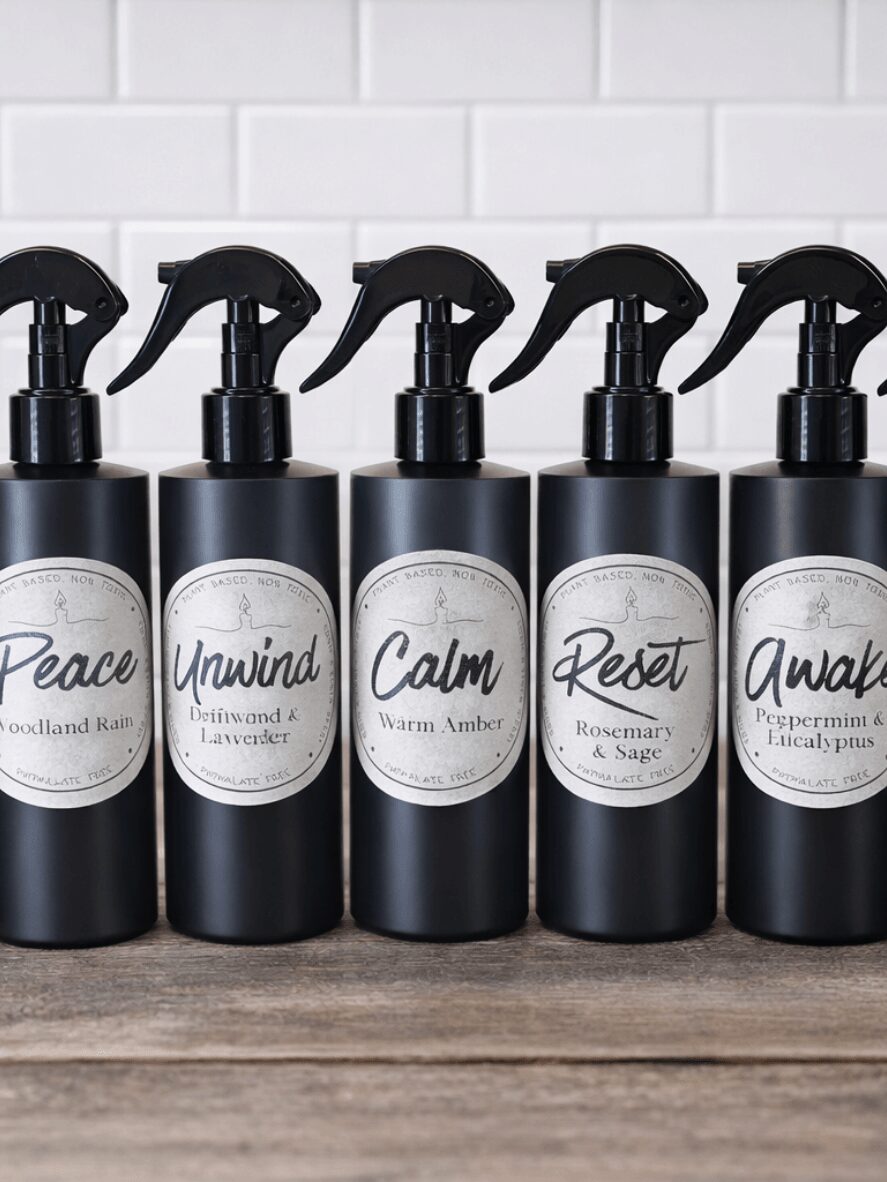 Five black spray bottles with white labels and black spray nozzles, labeled Peace, Unwind, Calm, Reset, and Awake, lined up against a white tiled background.