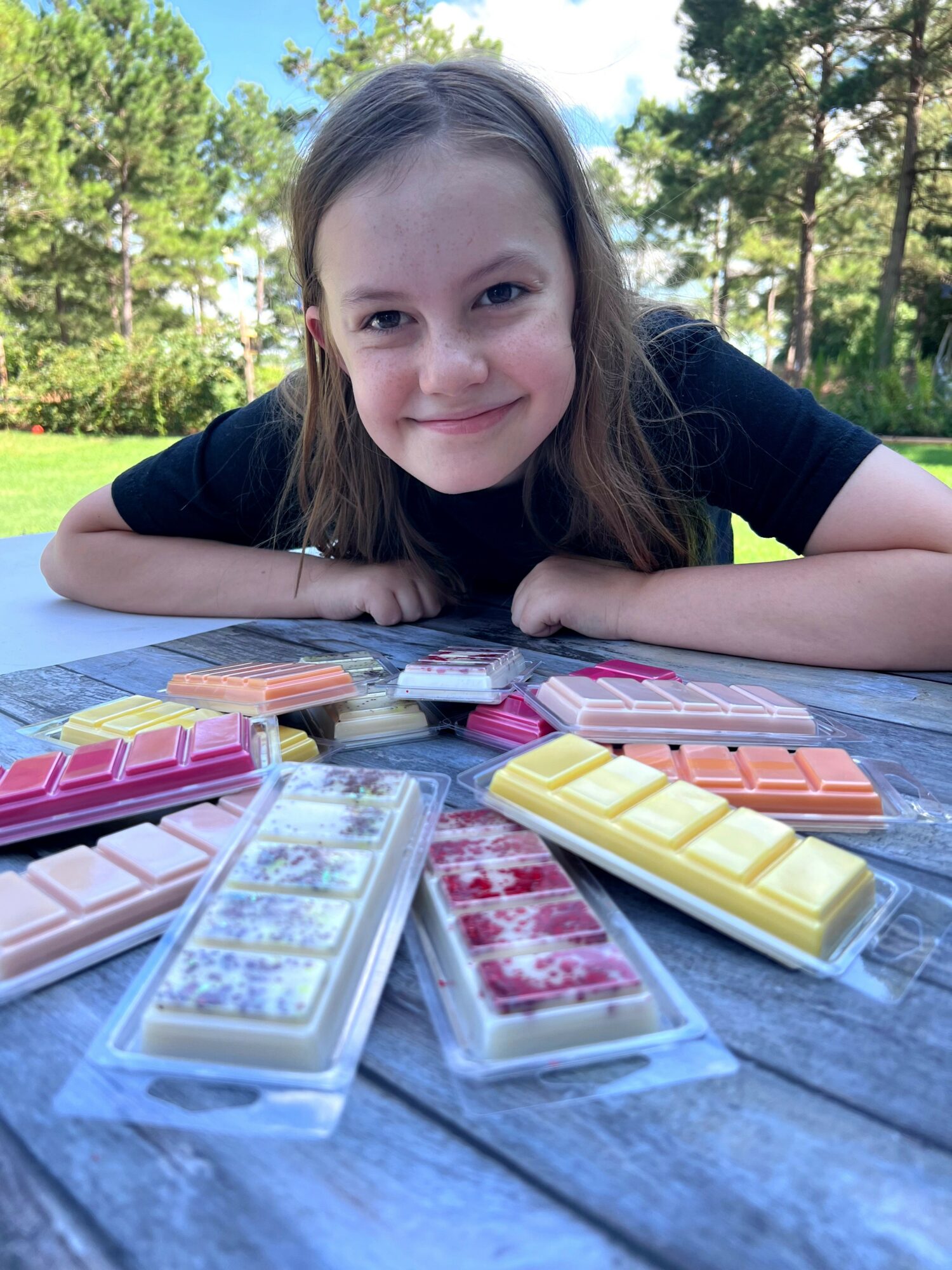 Girl lying on a table outdoors with colorful dominoes, smiling, trees and sky in background.